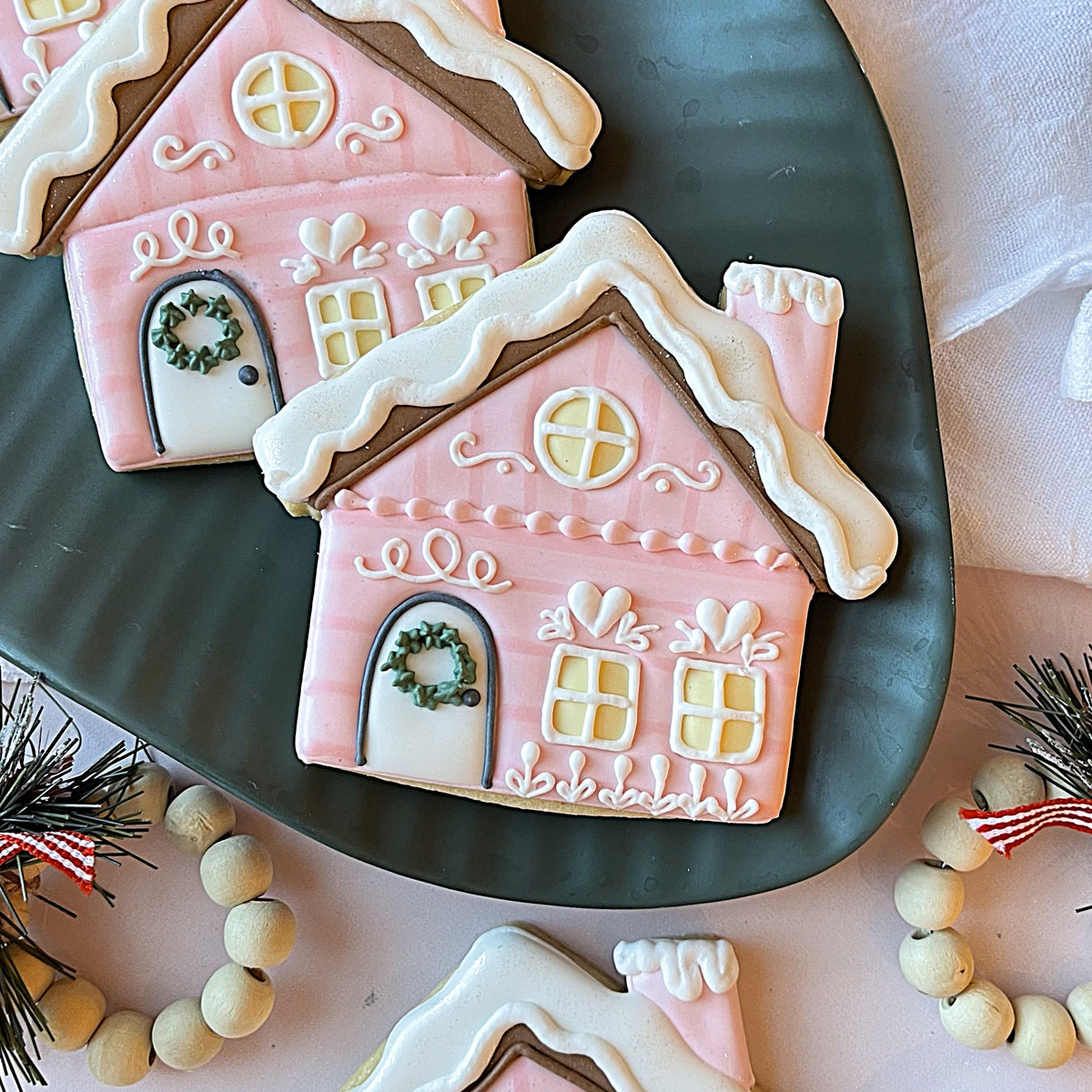 Pink gingerbread house cookies with white icing roofs and green wreaths made using snowy cabin flour box club cookie cutter