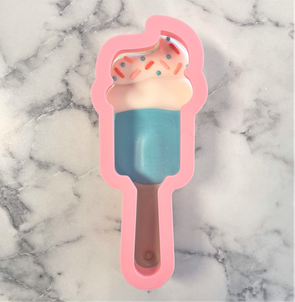 Pink silicone spatula cookie cutter mold with blue frozen center and pink frosting topped with colorful sprinkles