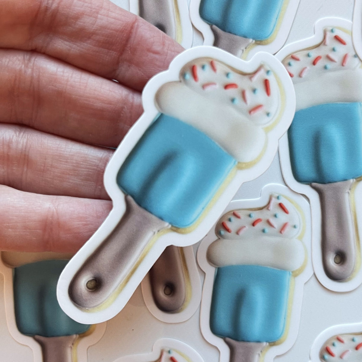 A blue popsicle-shaped spatula baking sticker with white frosting, colorful sprinkles, and a brown stick
