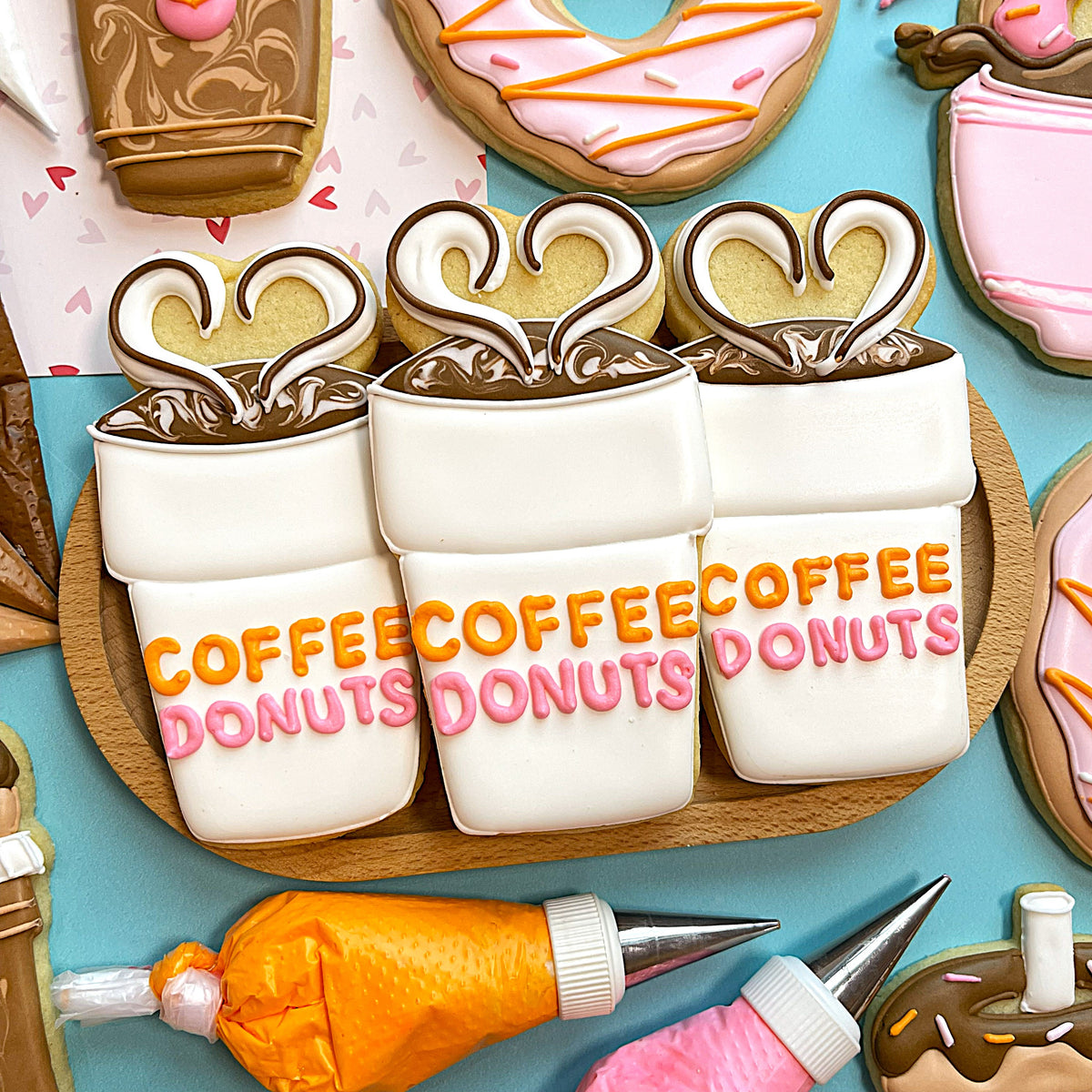 Three white frosted sugar cookies shaped like coffee cups with orange and pink icing spelling coffee donuts for donut online class recording