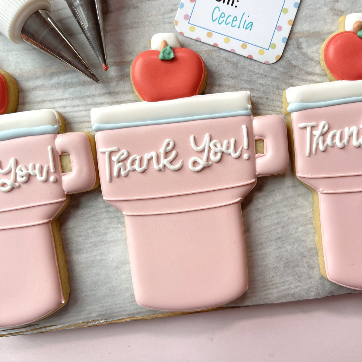 Pink frosted sugar cookie shaped like a mug with white thank you icing and red apple decoration made with teacher tumbler cookie cutter