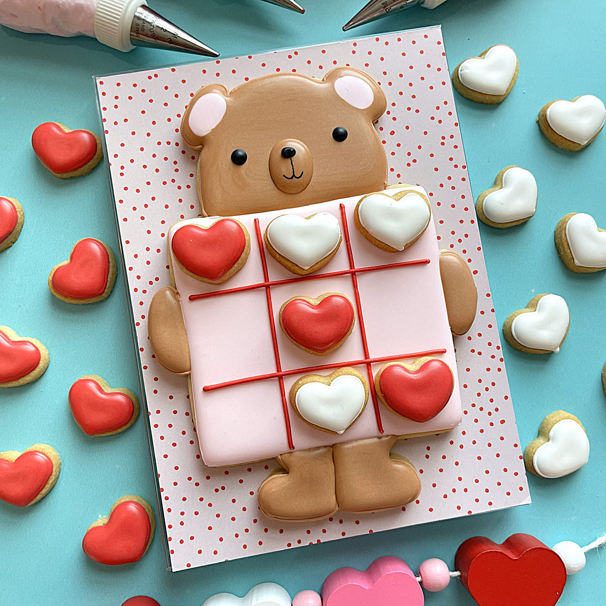 A charming brown bear-shaped cookie holding a tic tac toe game of red and white heart cookies decorated with icing