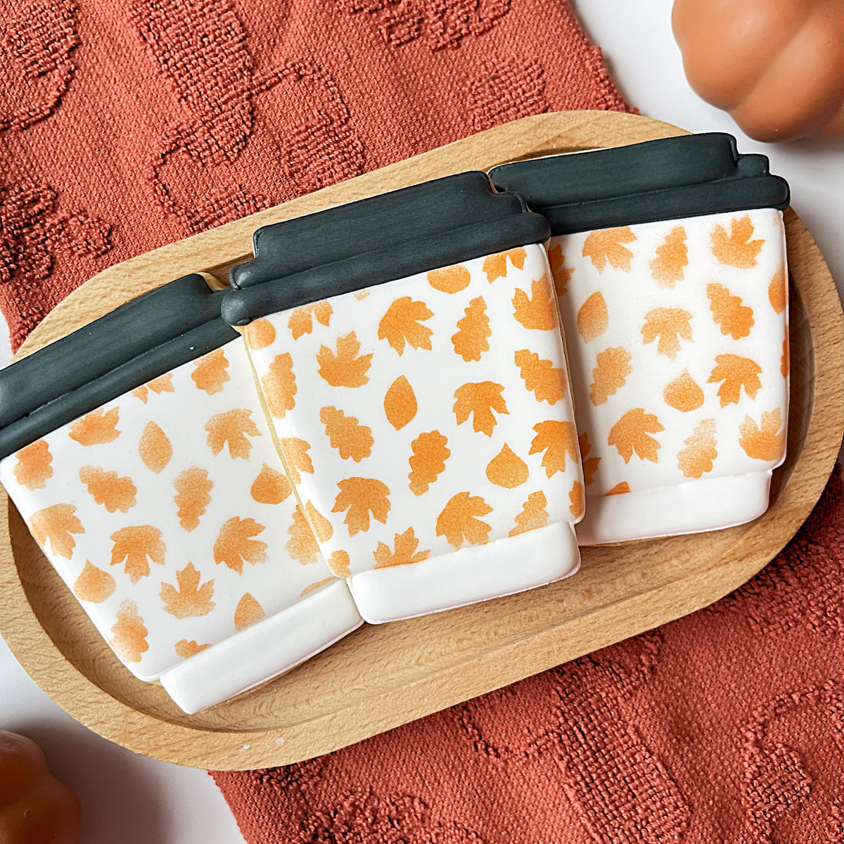 Three white ceramic coffee cup shaped cookies with orange leaf patterns and black lids on a wooden tray from travel coffee cup flour box club cookie cutter