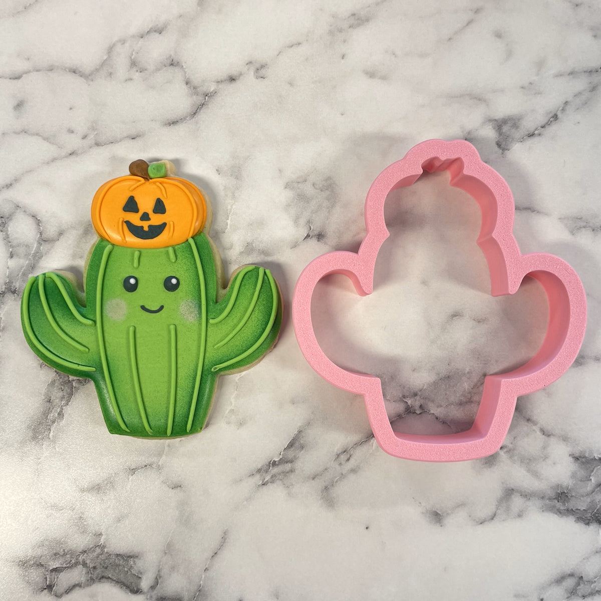 Green cactus shaped cookie with smiling face and orange pumpkin on top next to pink silicone mold for western cactus cookie cutter