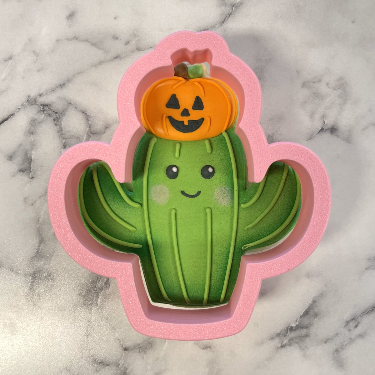 Pink glittery silicone cookie cutter shaped like a green cactus with black eyes and smiling orange pumpkin top