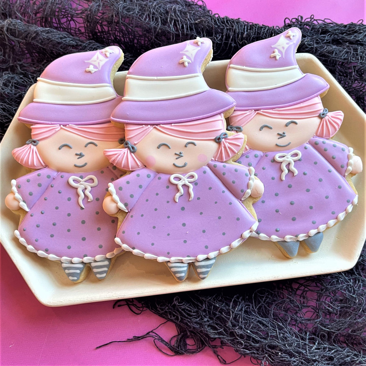 Three intricately decorated purple witch cookies with pink hair and white ribbon bows for cute witch online decorating class recording