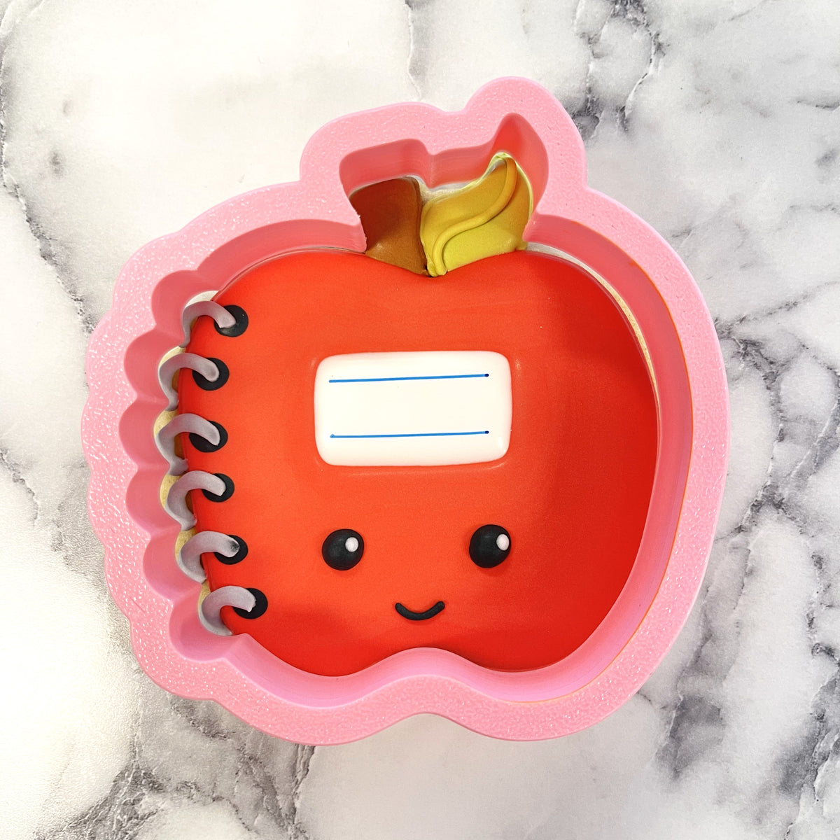 A pink silicone apple-shaped cookie cutter with a red smiling face interior and yellow stem for apple notebook cookies