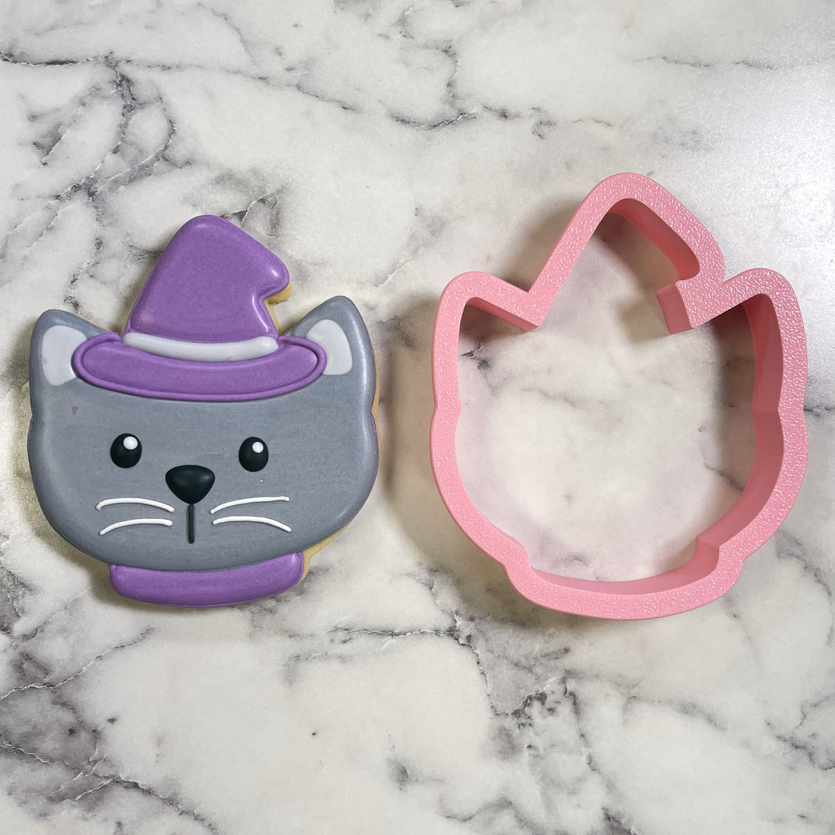 A cute chubby cat witch cookie cutter shaping a glossy purple and gray witch cat cookie made of pink silicone