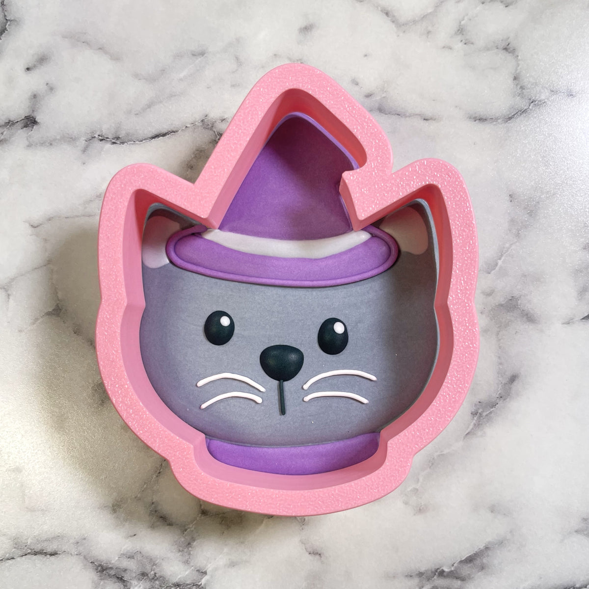 Cute chubby cat witch cookie cutter in pink silicone with purple witch hat and gray face details for baking