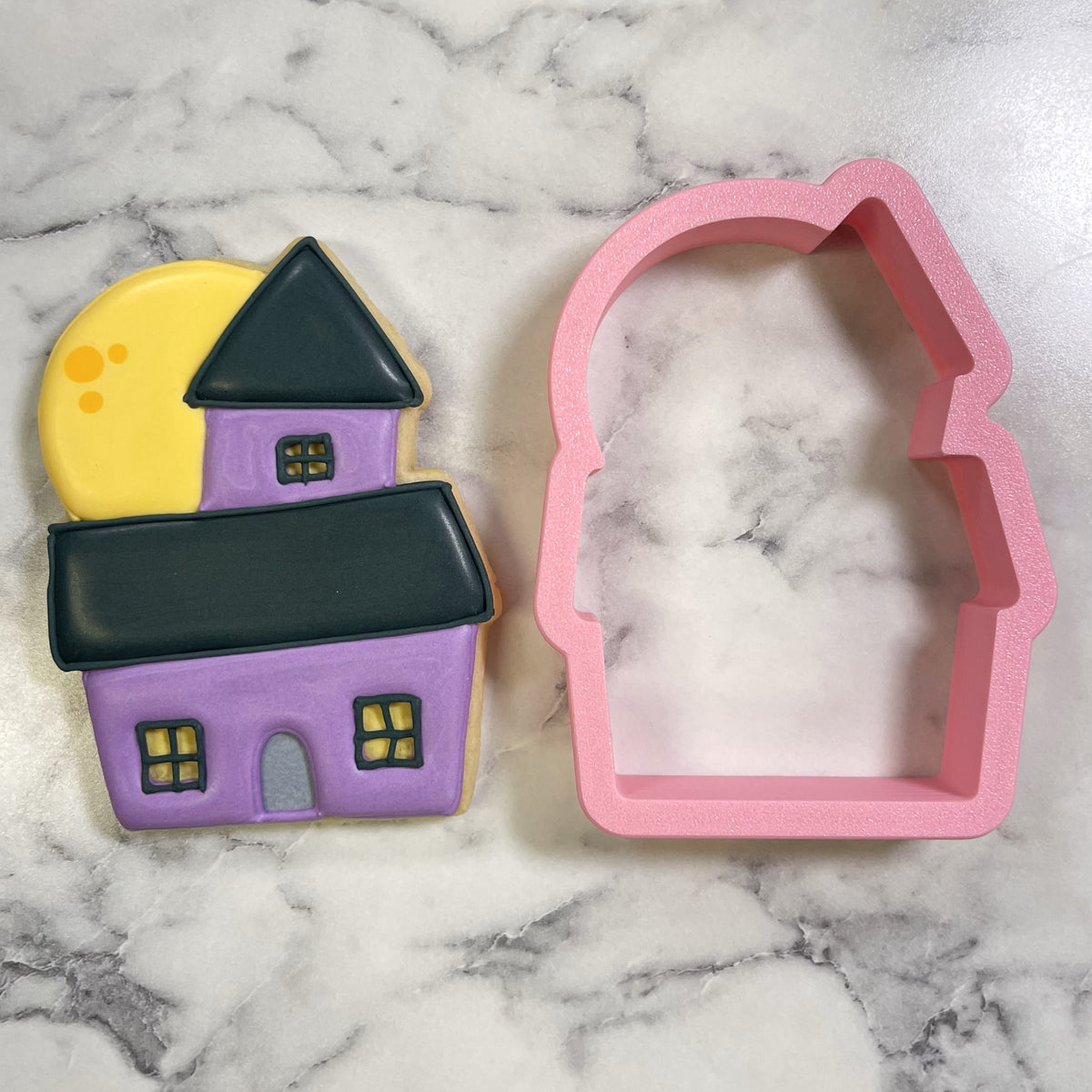 Cute chubby haunted house cookie cutter pink silicone next to purple and black decorated haunted house sugar cookie