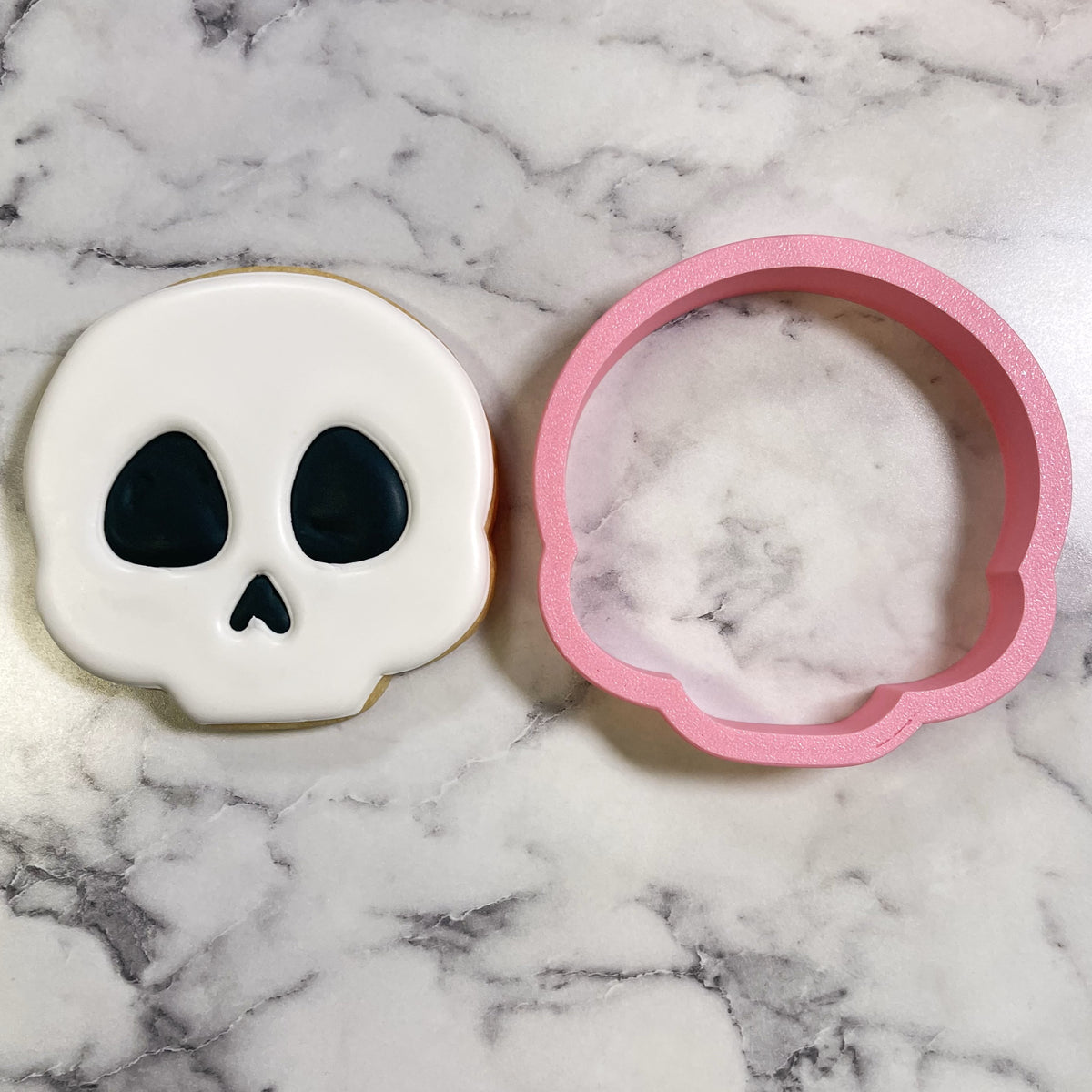 Cute chubby skull cookie cutter in pink plastic with smooth texture next to a white frosted sugar skull cookie
