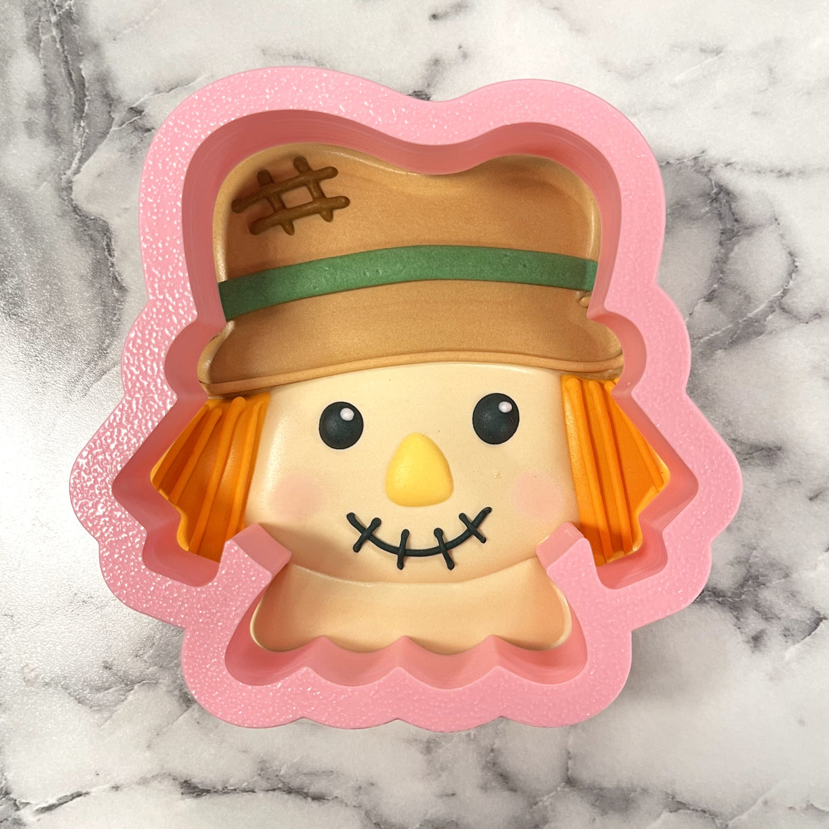 Pink silicone mold shaped like a cute scarecrow face with tan hat green band and stitched mouth for cookie cutter use
