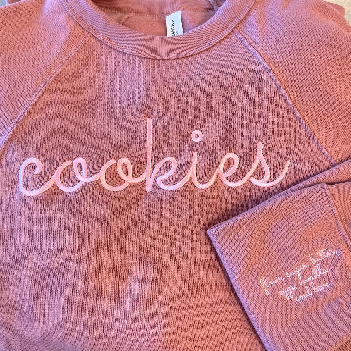 A soft rose-colored Cookies embroidered sweatshirt in mauve with cursive script on chest and matching pocket detail