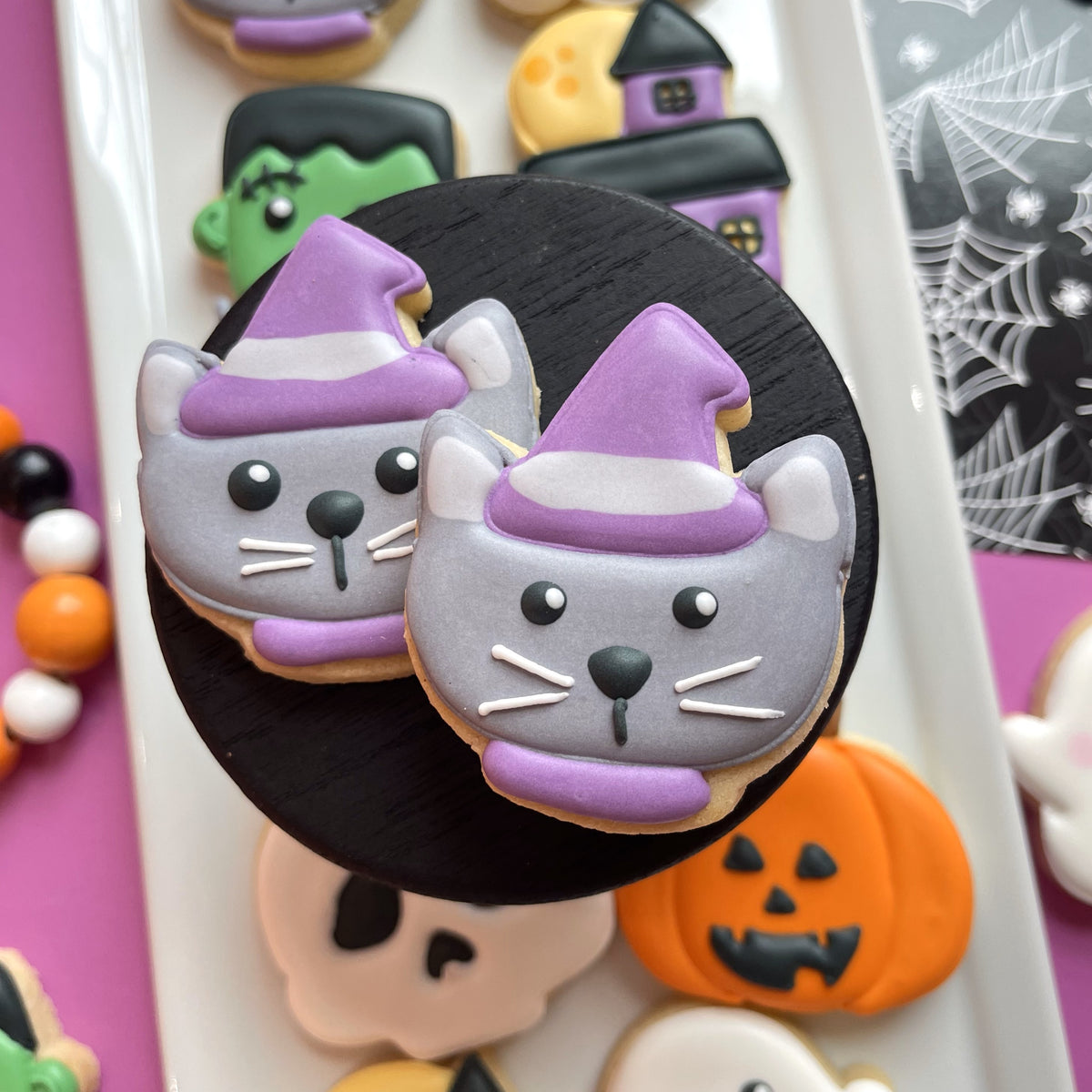 Two gray sugar cookies shaped like cats wearing purple witch hats with white trim from mini cute chubby halloween cookie cutter bundle