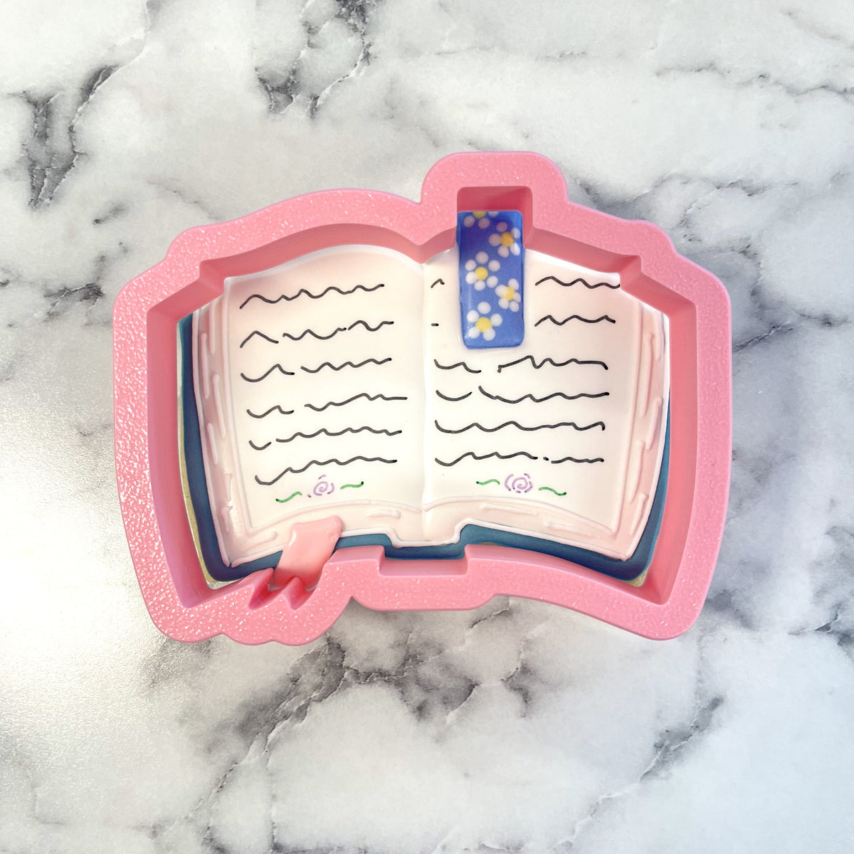 Pink book-shaped cookie cutter with blue floral bookmark and pink ribbon detail from bookish cookie cutter bundle with stencil