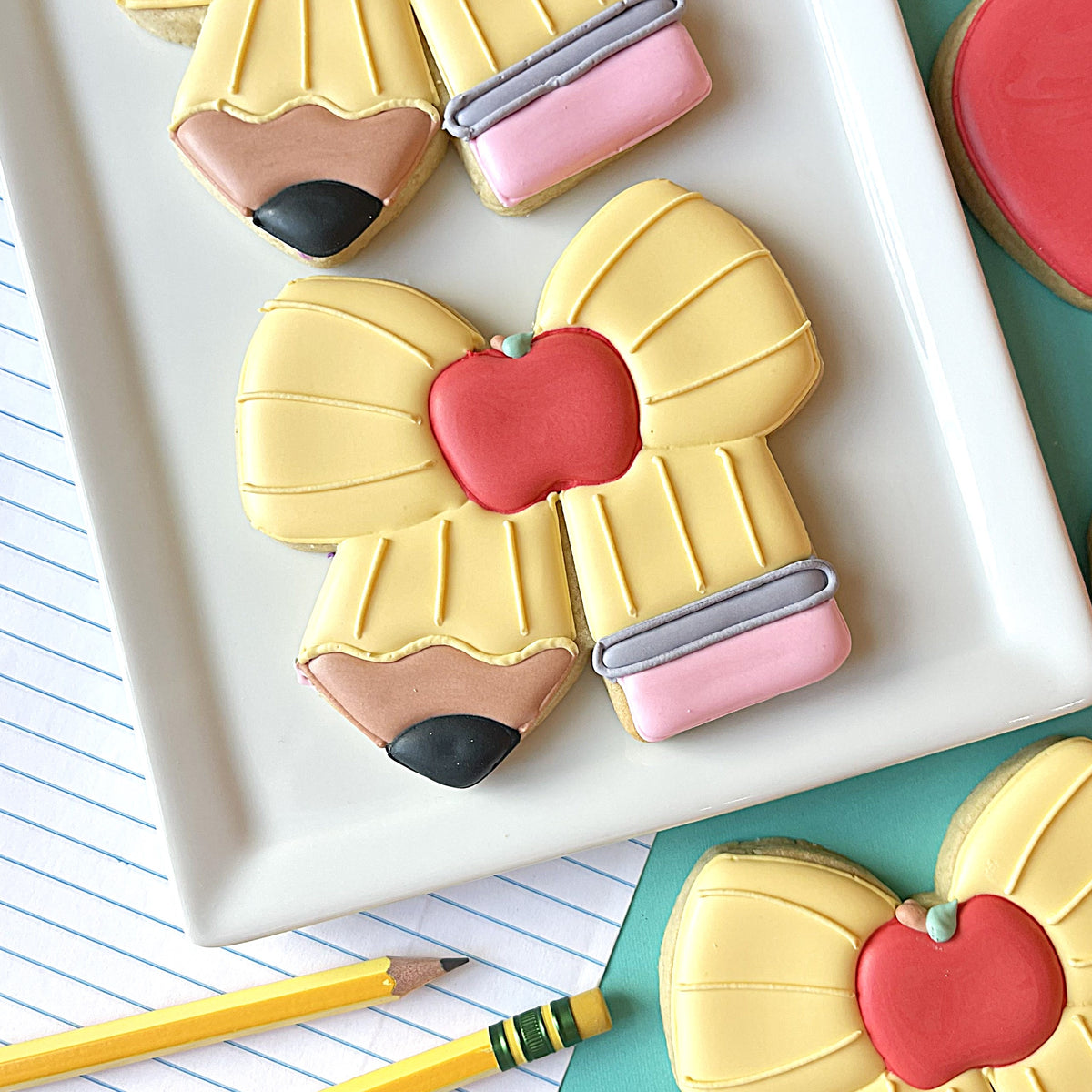 Yellow pencil bow cookie cutter shaped cookie with smooth icing and a red apple center decoration