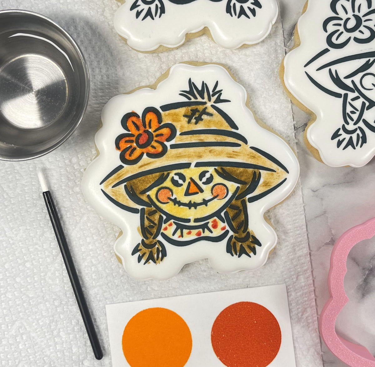 A hand-decorated scarecrow girl sugar cookie with black outlines and an orange flower made using a PYO cookie cutter bundle