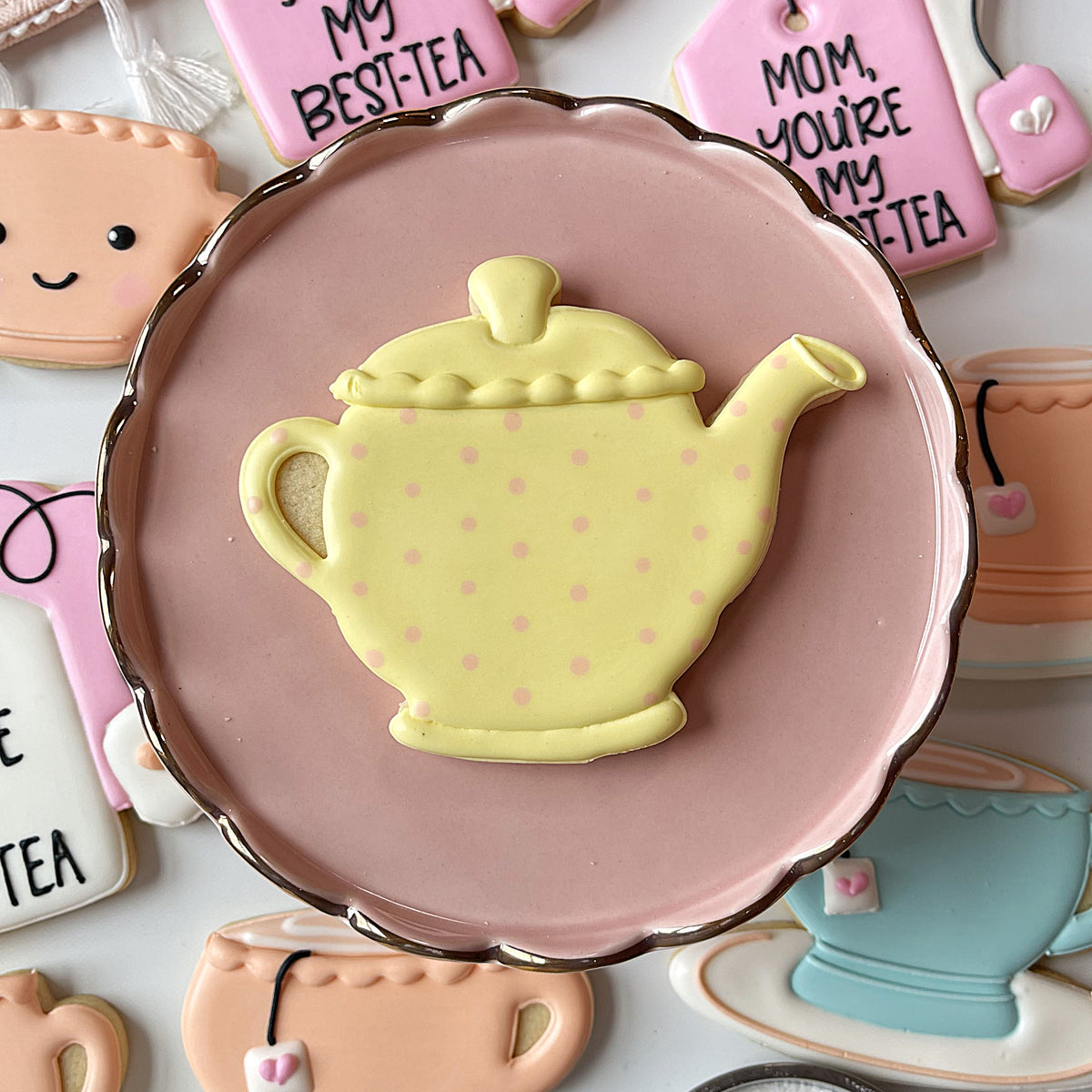 Pale yellow teapot-shaped cookie with pink polka dots and gold trim on soft pink plate from teatime cookie cutter bundle