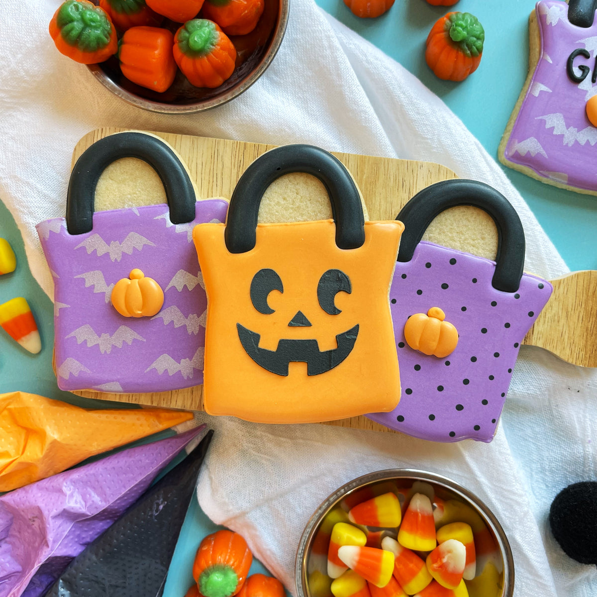 Three halloween-themed sugar cookies shaped like handbags with an orange jack-o-lantern and purple bat patterns treat bag flour box club cookie cutter