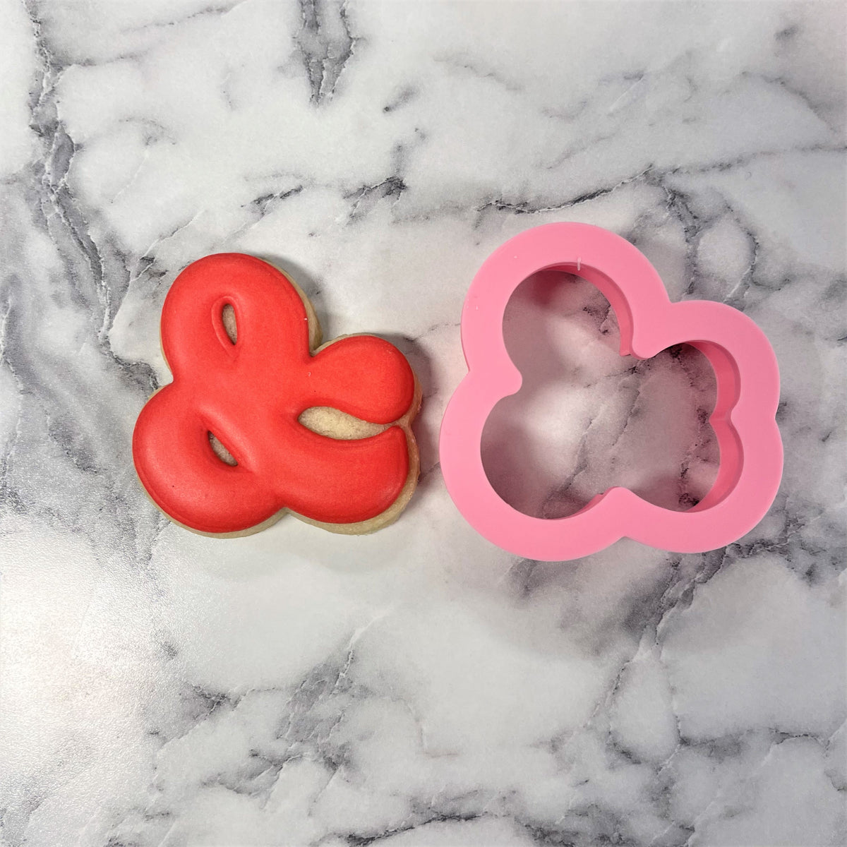 A vibrant red glossy frosted mini ampersand cookie next to a pink silicone cutter on a marble surface
