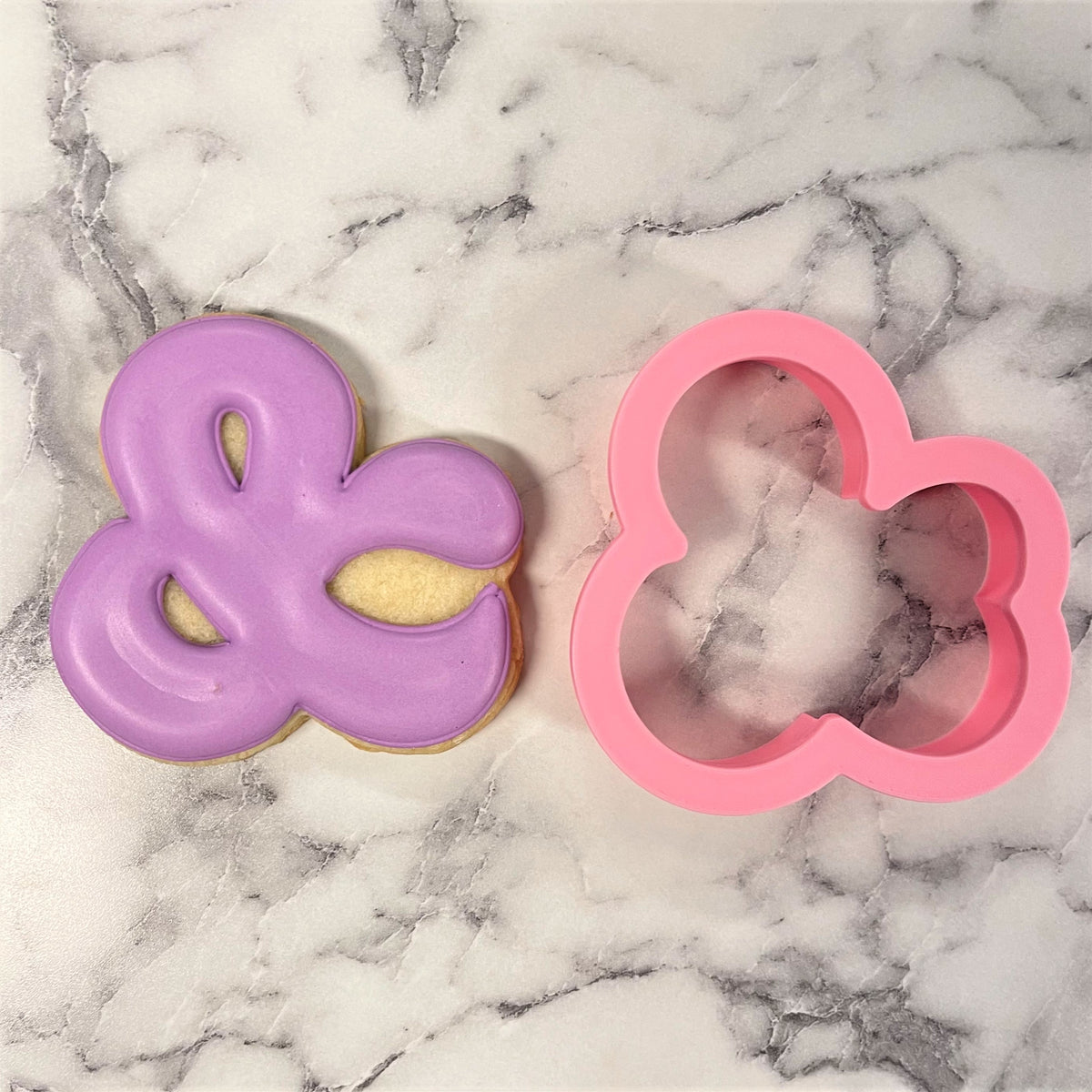 Ampersand cookie cutter pink silicone four petaled flower shape beside matching purple iced cookie