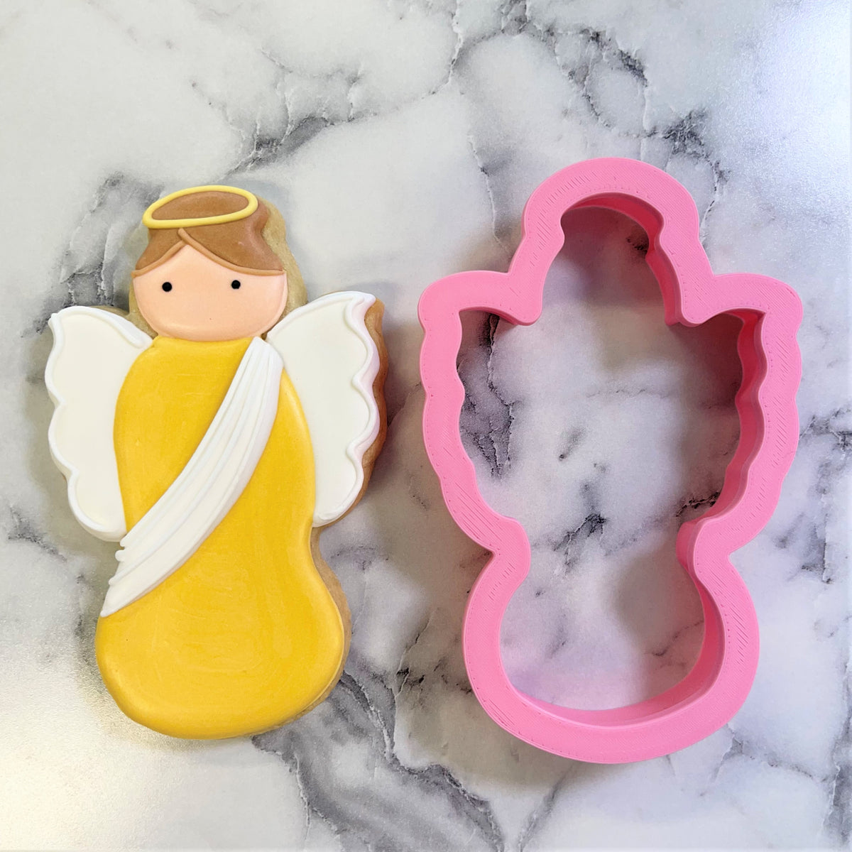 A pink silicone christmas angel cookie cutter next to a yellow and white frosted angel cookie