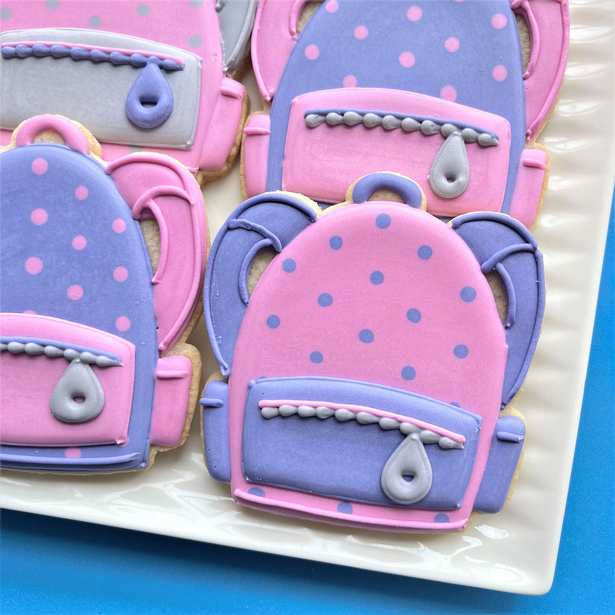 Pink and purple polka-dotted backpack-shaped sugar cookies with gray icing made using a backpack cookie cutter