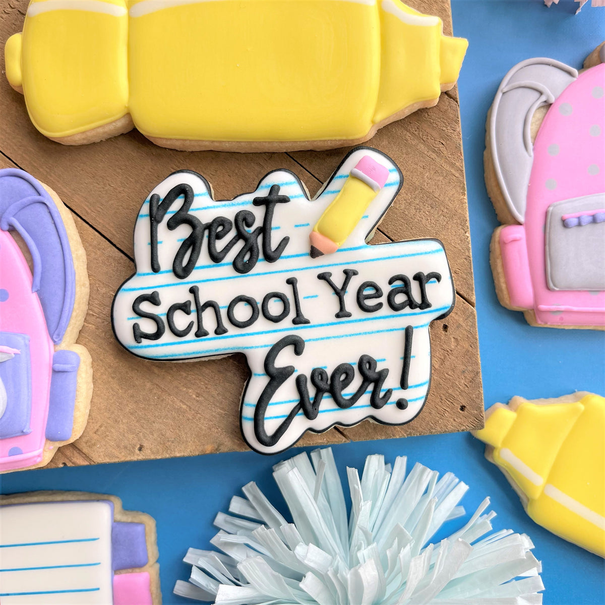 A white sugar cookie shaped like a notebook page with best school year ever icing and a pencil detail featuring the best school year ever plaque cookie cutter
