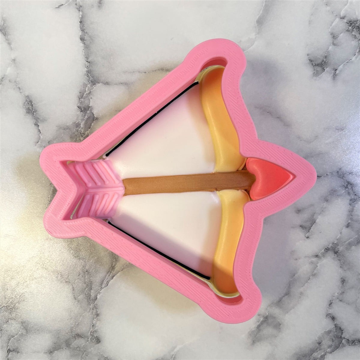 Bow and arrow cookie cutter made of pink plastic with yellow string brown handle and red arrowhead for baking