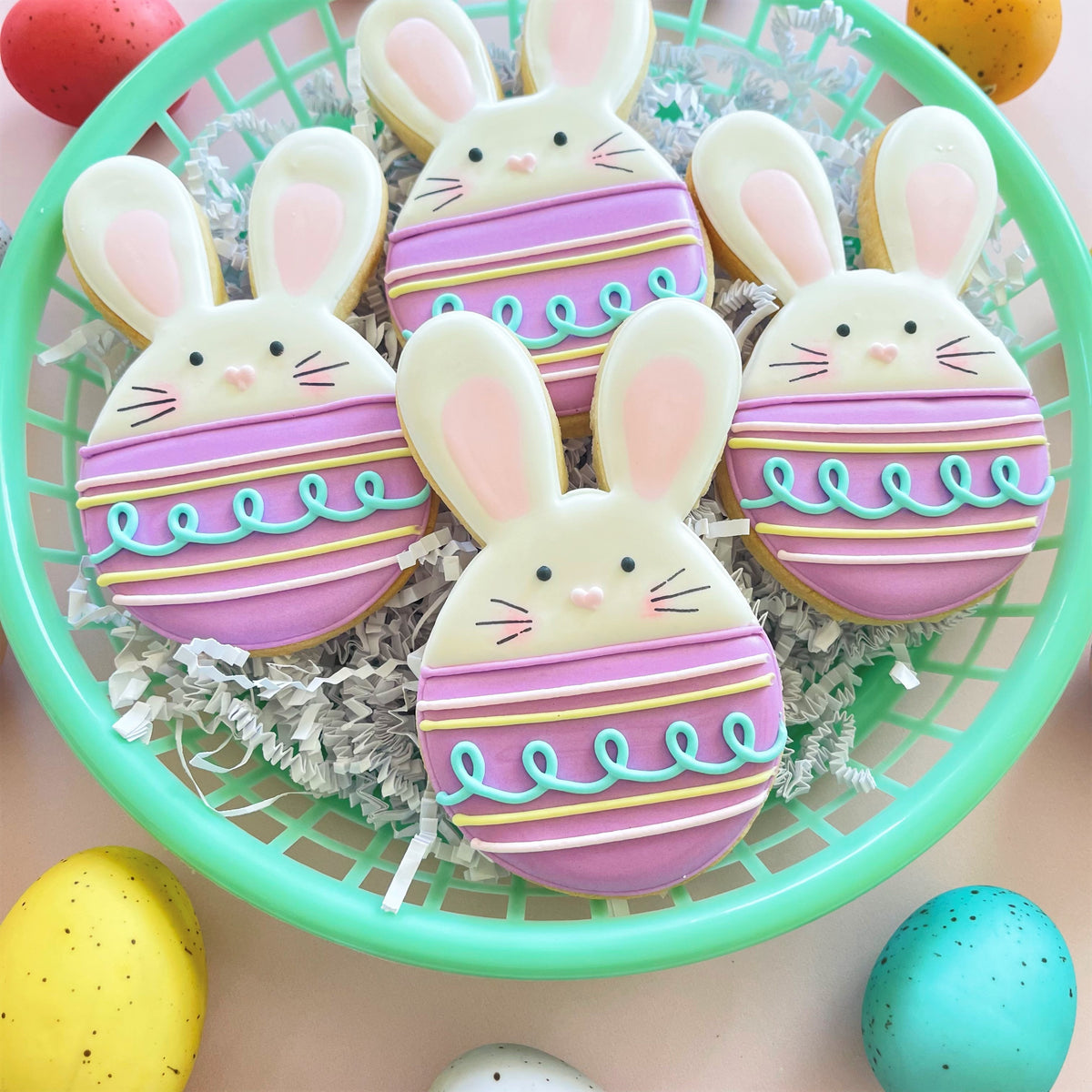 Bunny egg cookie cutter shaped cookies in pastel pink and white with blue and yellow icing stripes in a mint basket