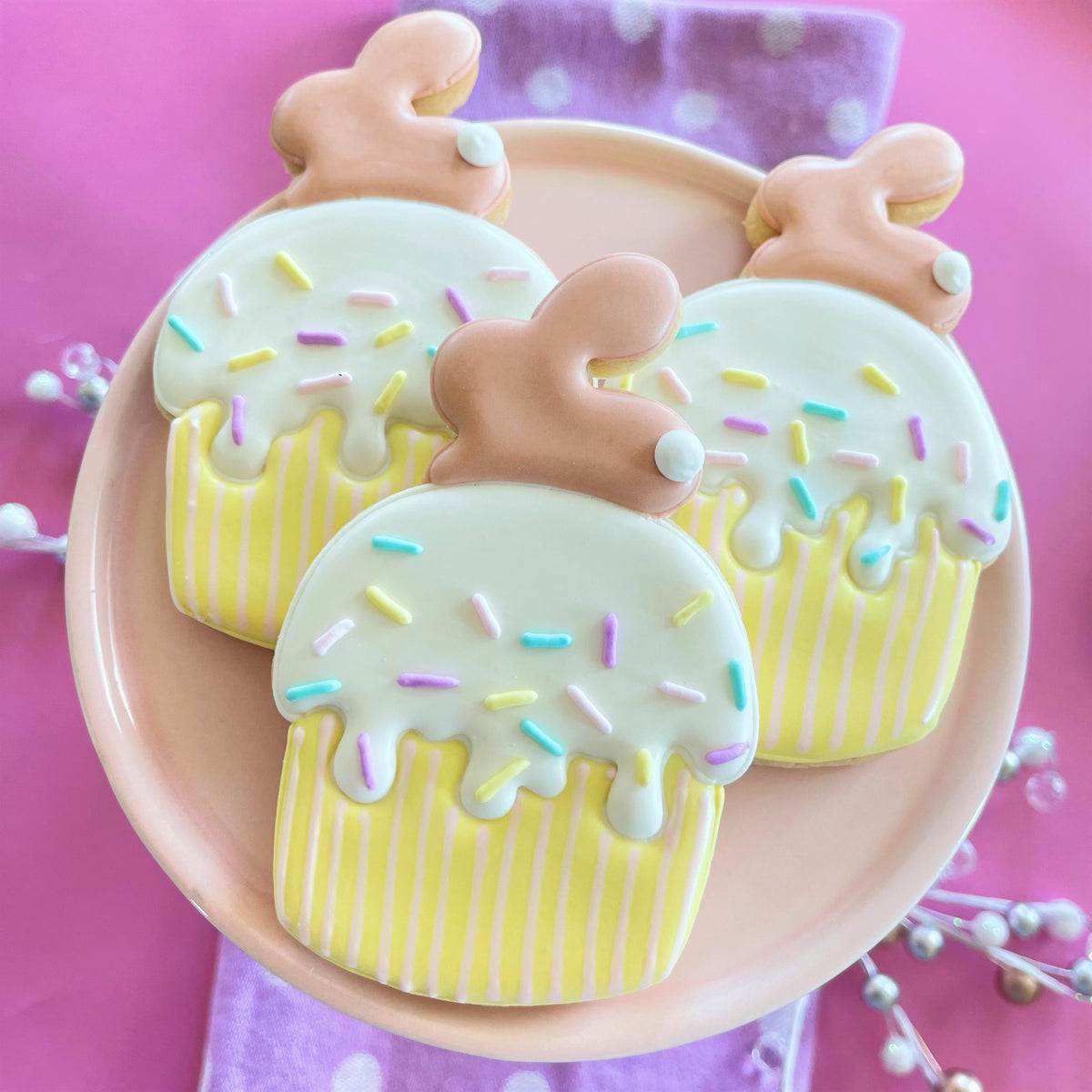 Pastel yellow cupcake-shaped cookies with white icing and sprinkles topped with tan bunny figurines made using bunny muffin cookie cutter