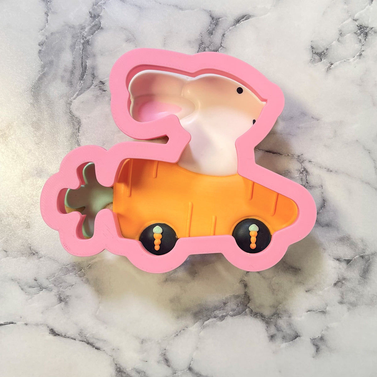 Pink silicone bunny shaped cookie cutter with carrot carriage and black wheels featuring orange details