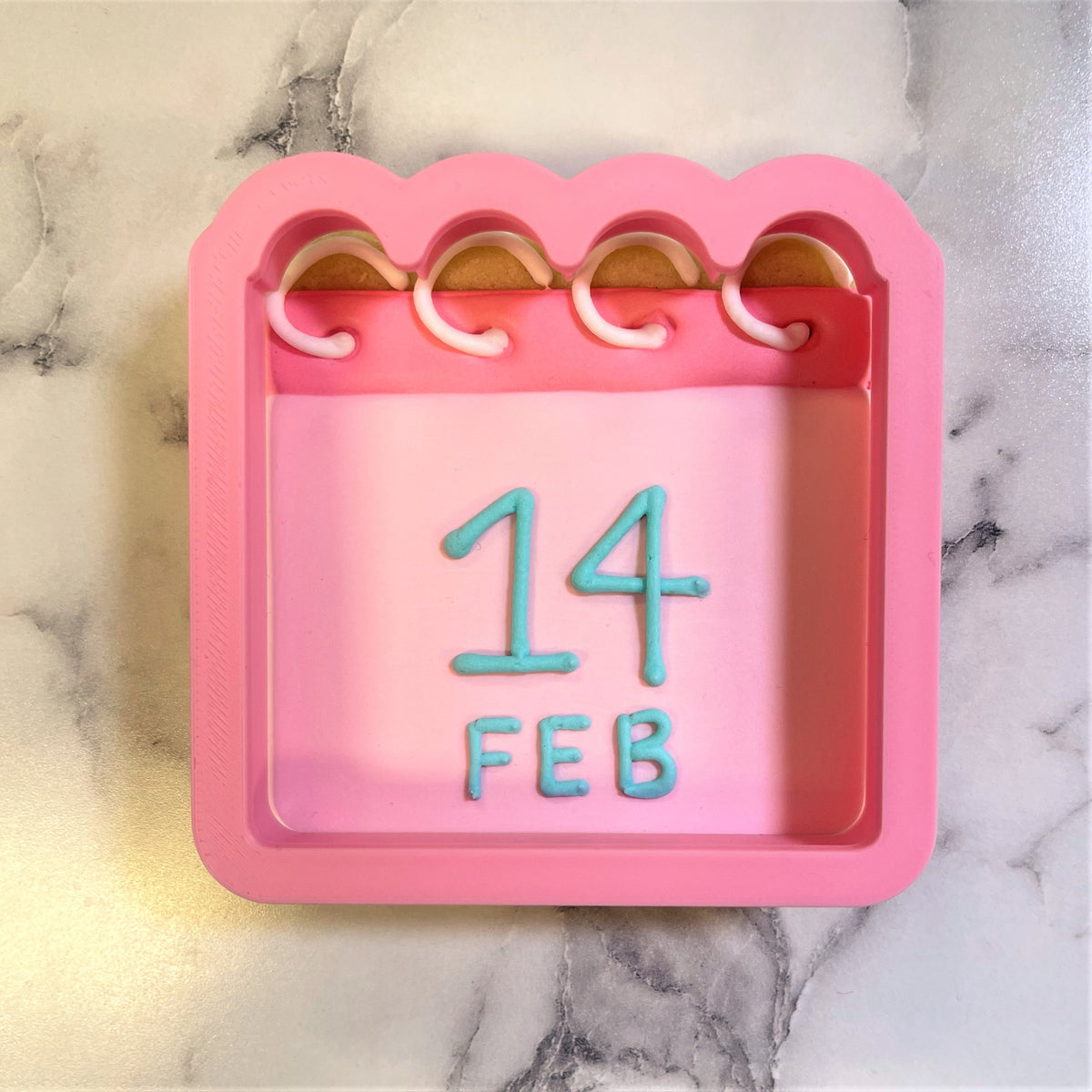 Pink silicone calendar cookie cutter mold with scalloped edges and light blue raised numbers reading 14 february