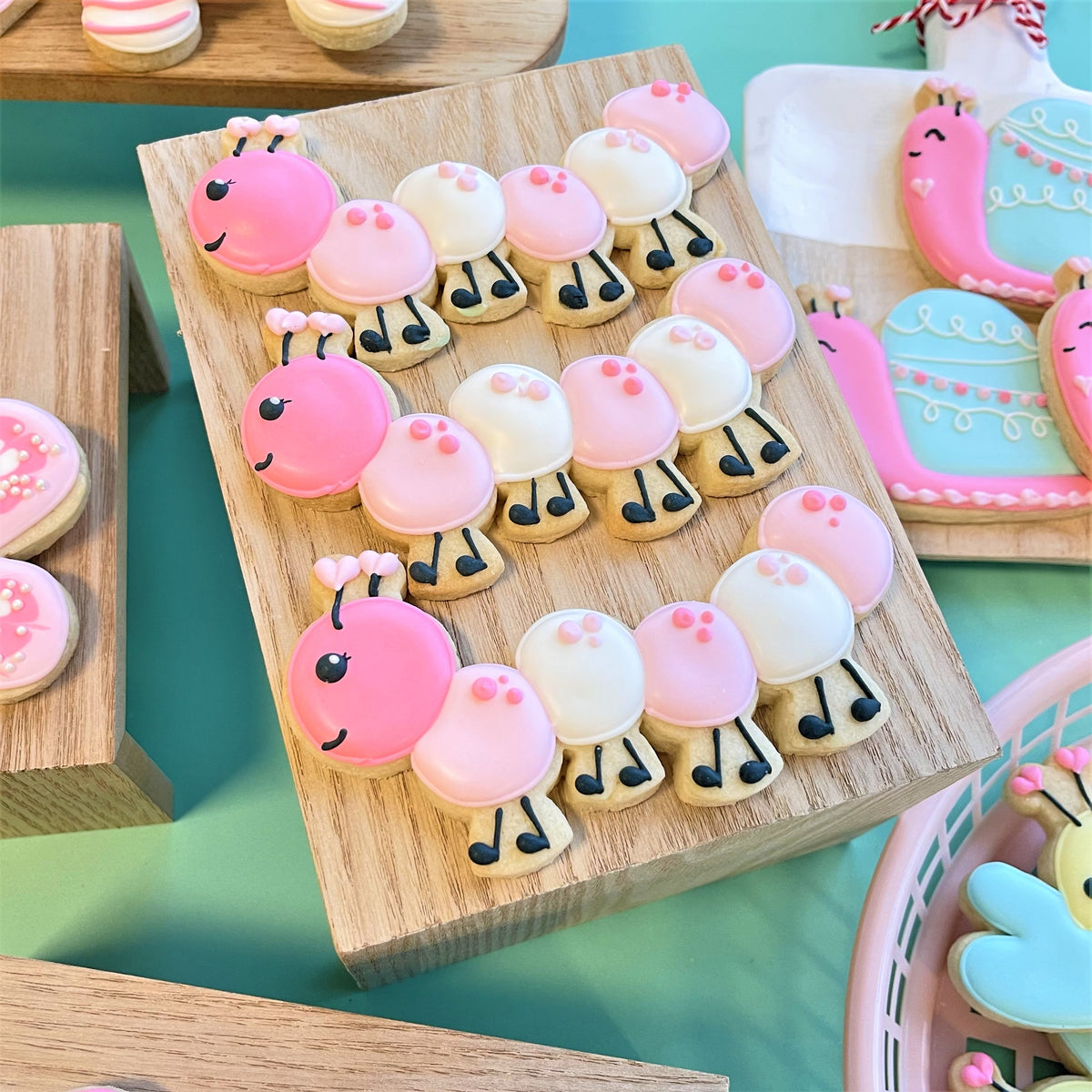 Pink and white frosted caterpillar-shaped cookies with black icing legs on a wooden board made with caterpillar cookie cutter
