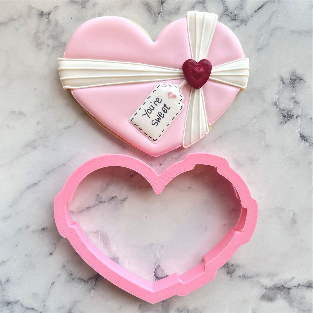 Pink heart-shaped metal cookie cutter with matching pink frosted cookie decorated with white ribbon and red heart charm