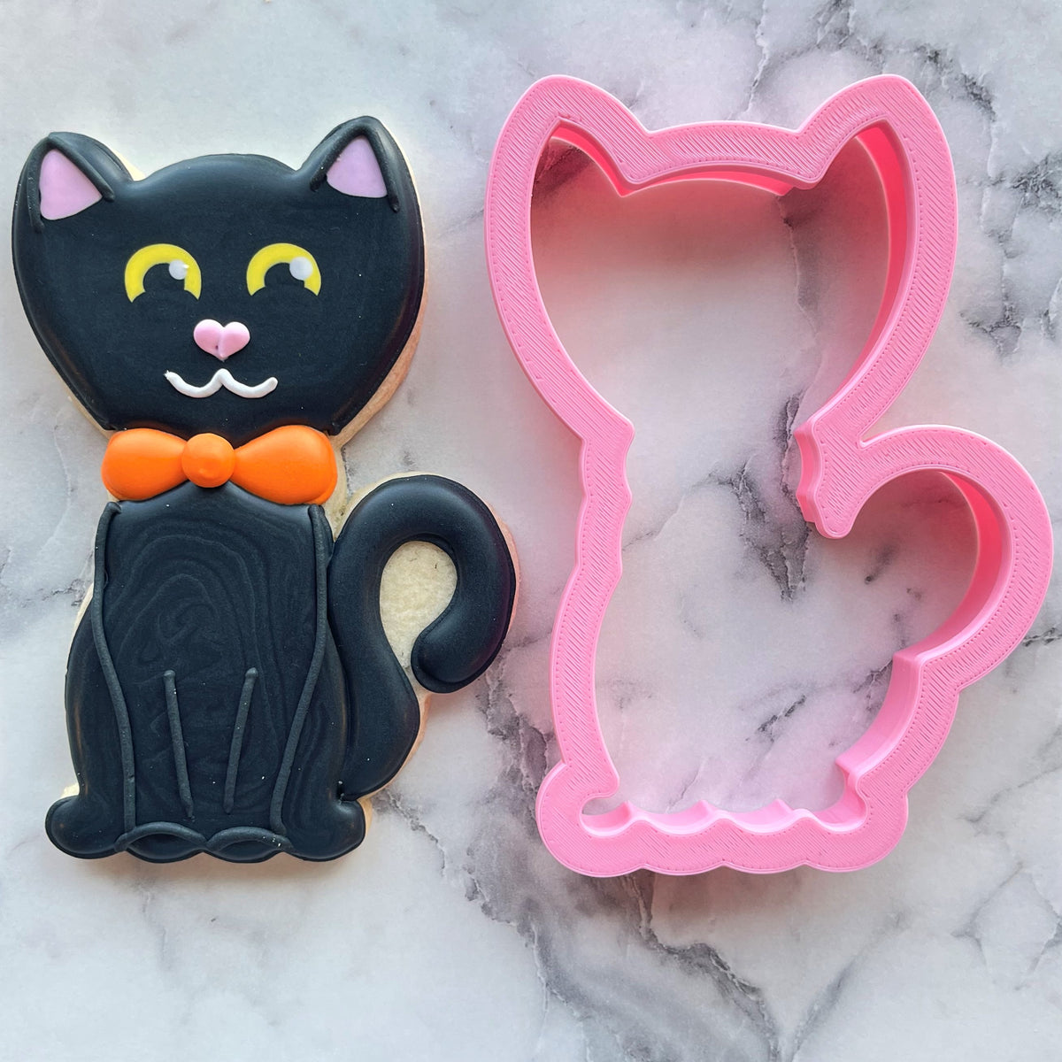 A black cat-shaped cookie decorated with yellow eyes pink nose and orange bow tie for classic black cat cookie cutter