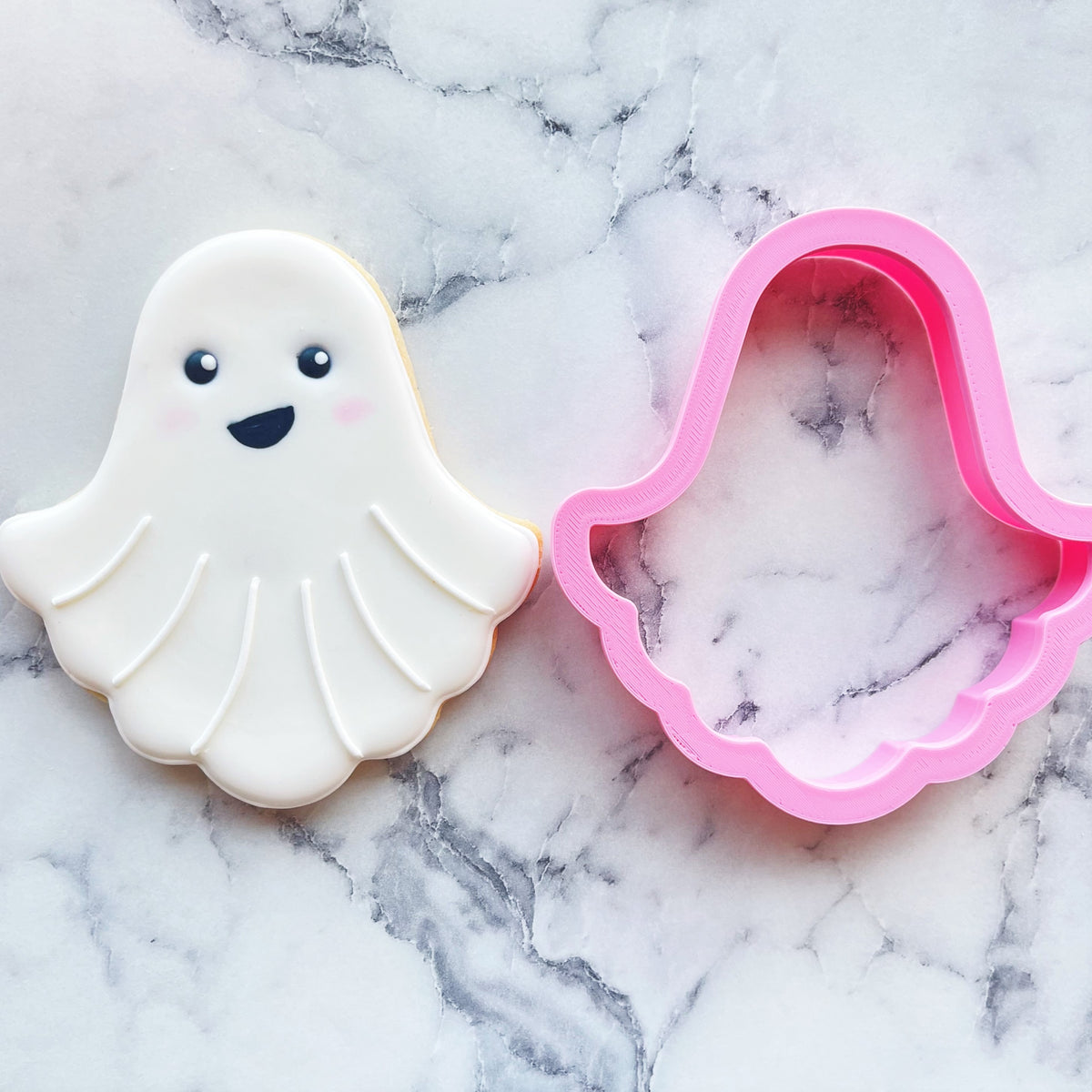 Classic ghost cookie cutter in pink with a glossy finish beside a white frosted smiling ghost cookie