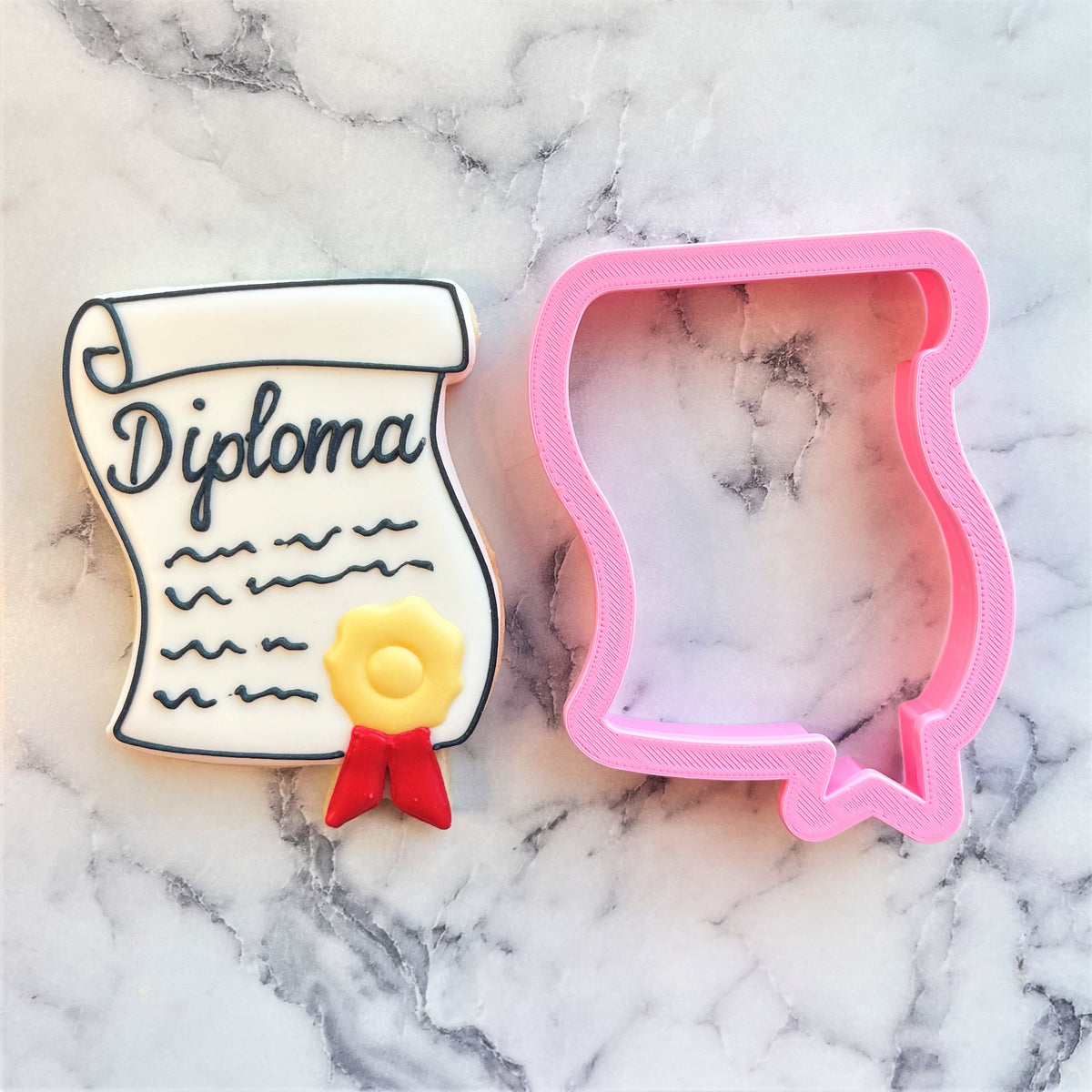 A pink plastic diploma with seal cookie cutter next to a white cookie decorated with black icing text and a yellow seal