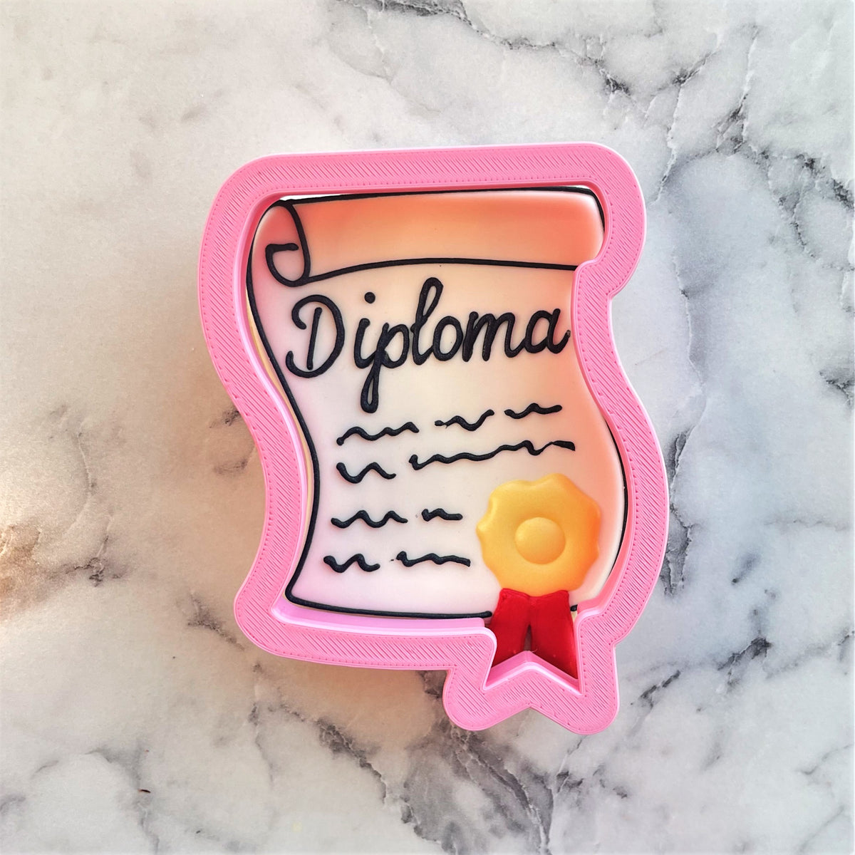 Pink plastic diploma with seal cookie cutter featuring black cursive lettering wavy lines and yellow seal with red ribbon