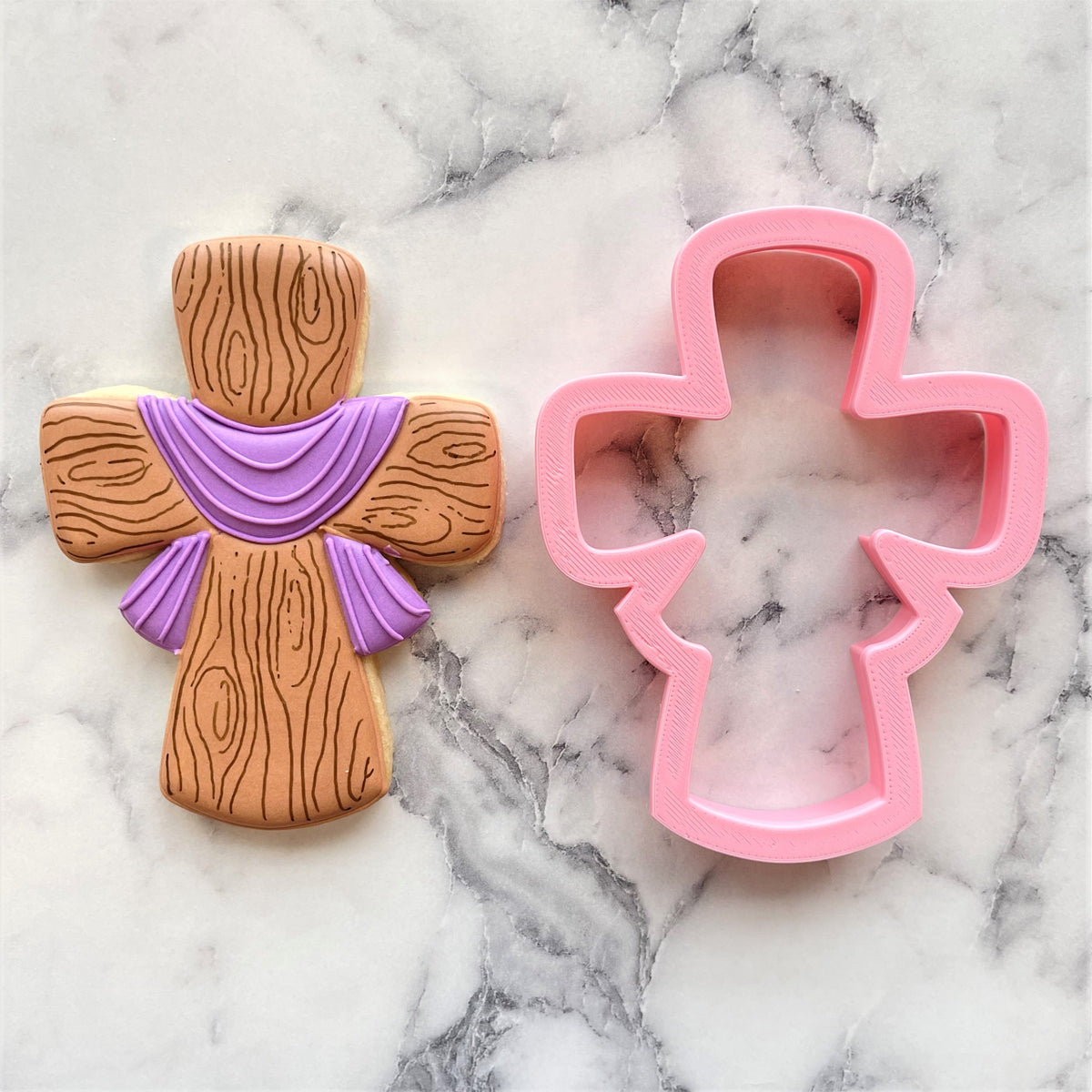 A wooden-textured cross-shaped cookie with a purple ribbon design and a pink silicone easter cross cookie cutter