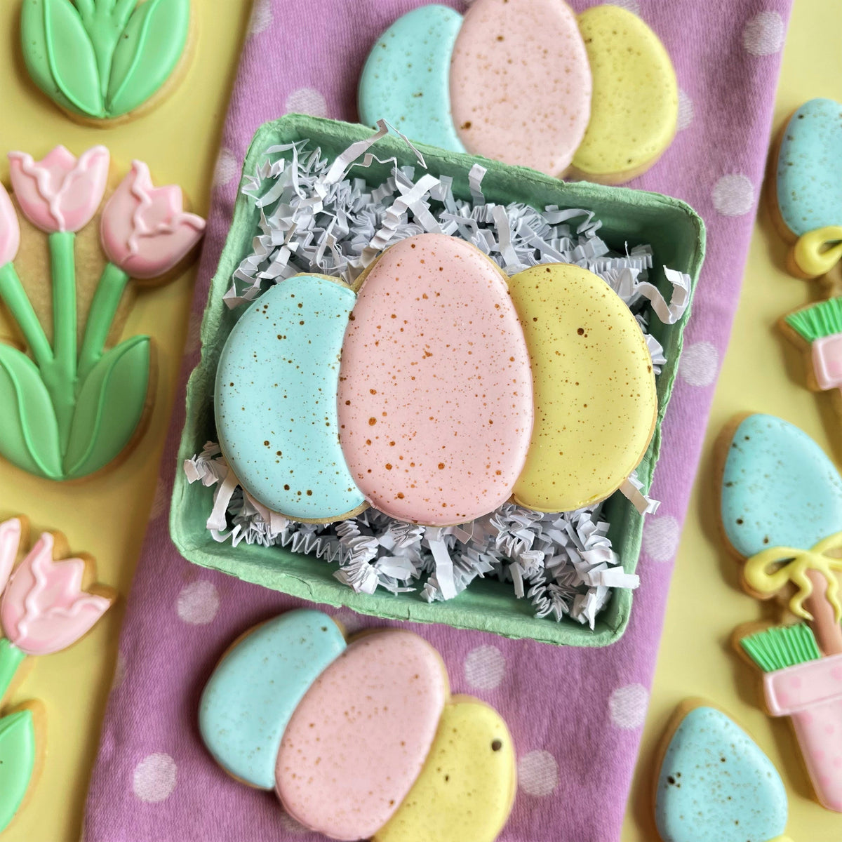 Pastel-colored egg-shaped sugar cookies with speckled icing in a green carton for rustic easter online decorating class recording