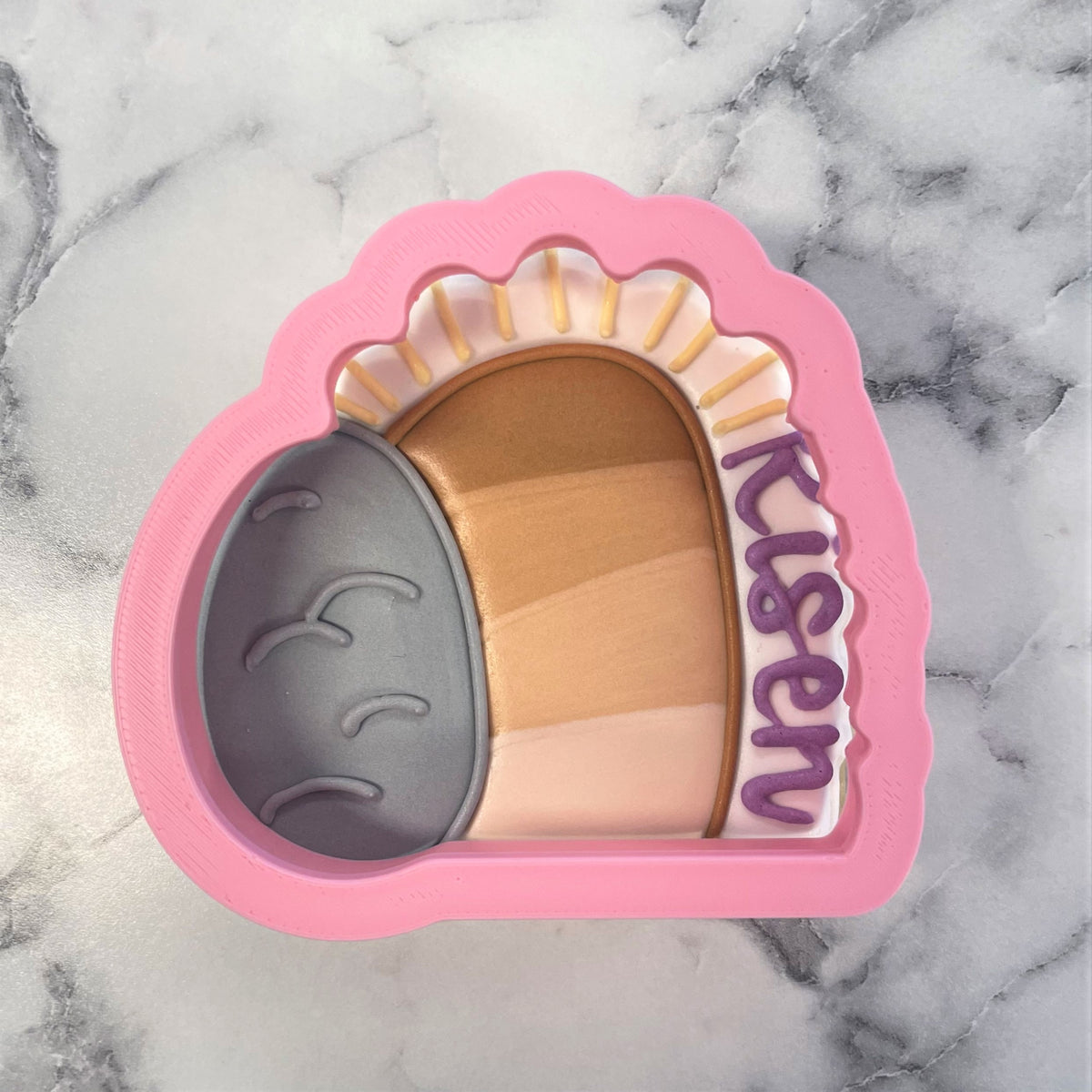Pink silicone empty tomb cookie cutter shaped like a smiling acorn with risen in purple lettering on the side