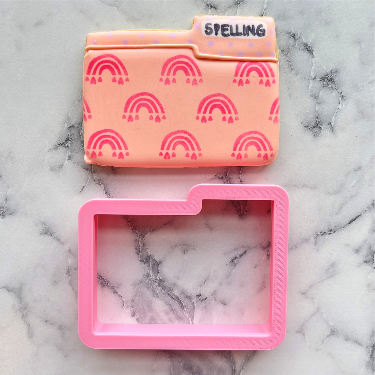 A pink silicone folder cookie cutter with a peach sugar cookie decorated with red rainbows and the word spelling