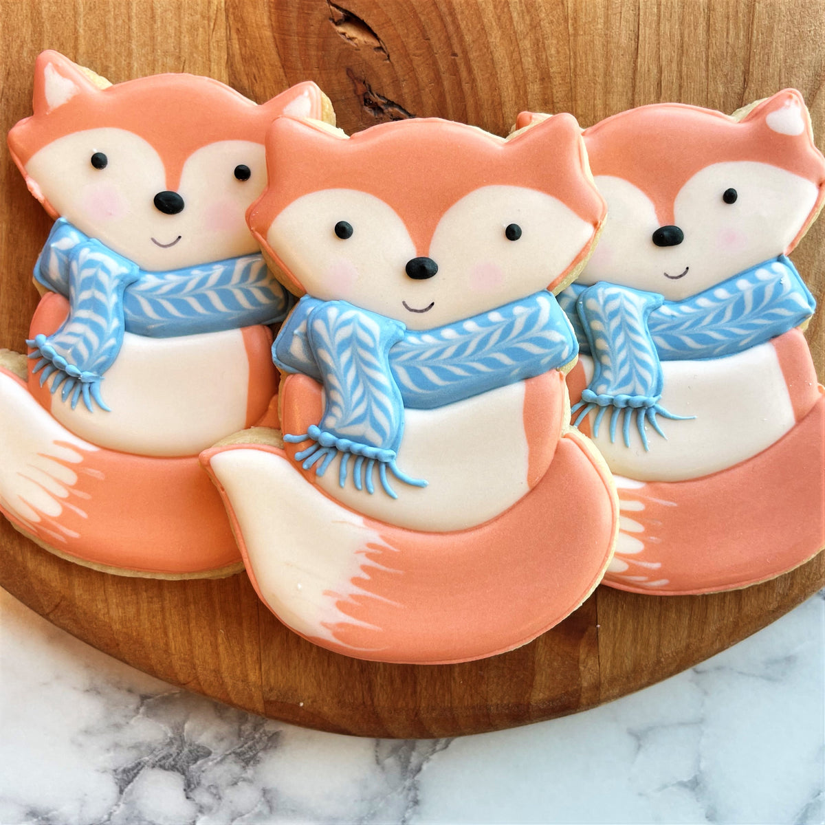 Cute orange fox-shaped sugar cookies with white accents and blue knitted scarf made using woodland fox cookie cutter