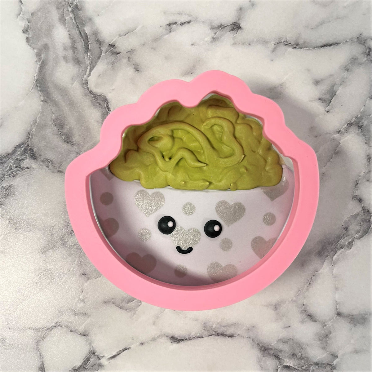 Pink silicone guacamole cookie cutter shaped like a cupcake with green brain filling and white base with silver hearts