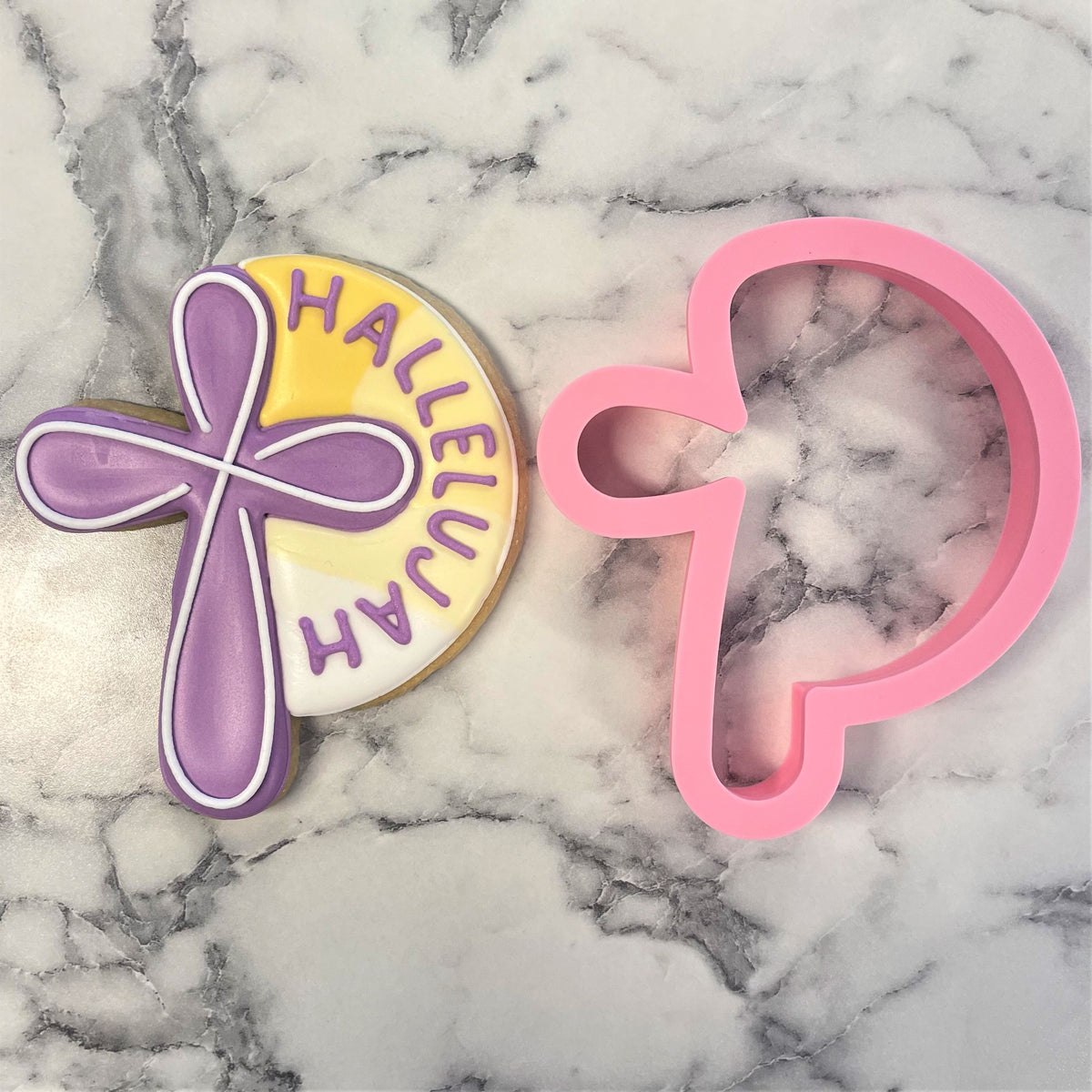 Pink heart-shaped hallelujah cross cookie cutter next to decorated cookie with purple cross on yellow background