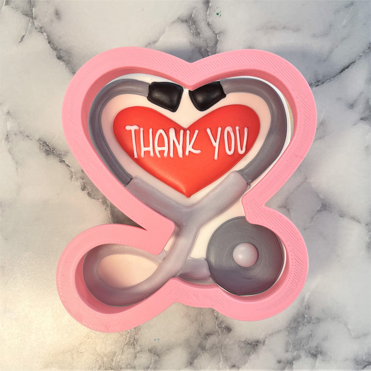 Pink heart-shaped stethoscope cookie cutter with red center reading thank you and black earpieces for baking