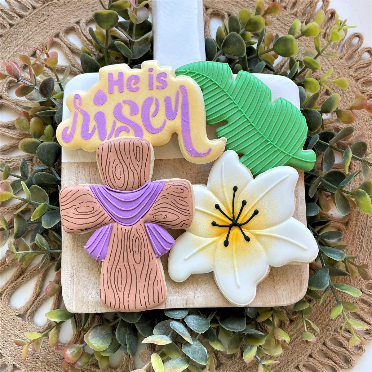 A decorative easter-themed cookie arrangement with a wooden textured cross purple accents flower leaves and he is risen icing using palm branch cookie cutter