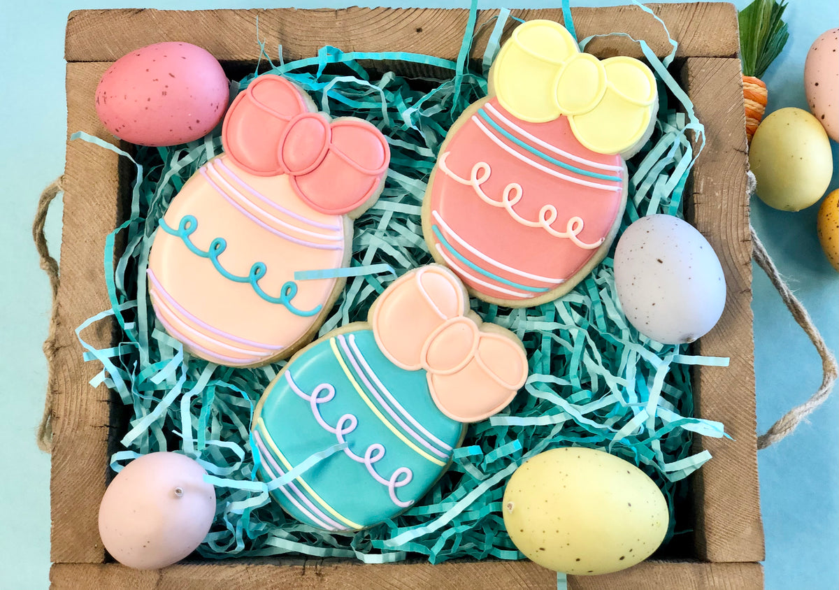 Pastel-colored easter egg-shaped sugar cookies with decorative bows and icing stripes in a wooden box with blue shredded paper