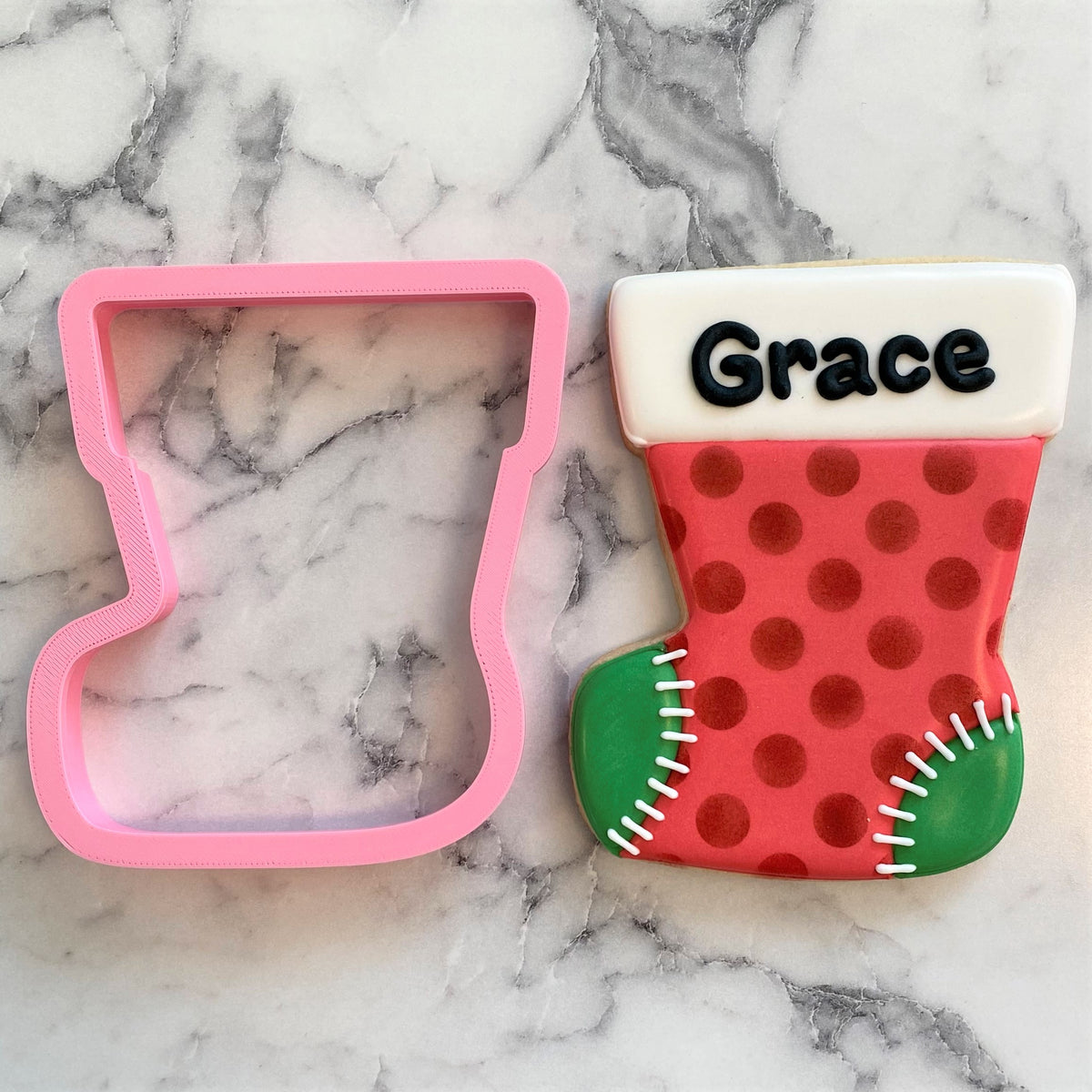 A pink plastic jumbo christmas stocking cookie cutter next to a decorated red and green sugar cookie with grace written in black icing